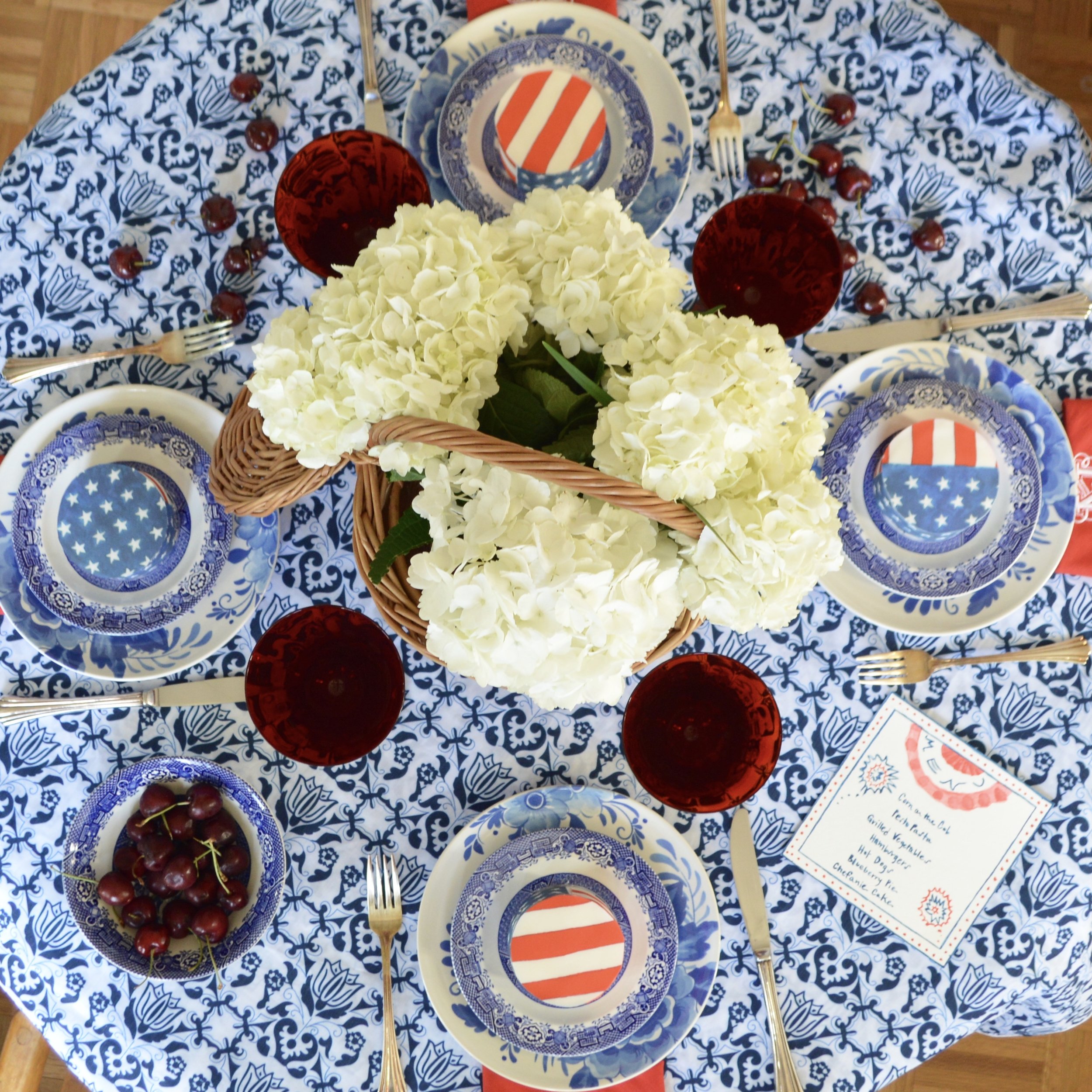 Four-Person July 4 Table with  Blue & White Tablecloth ,  Custom Embroidered Napkins , White Hydrangeas in  Wicker Basket , and Personalized Cakes with  Flag Chefanie Sheets