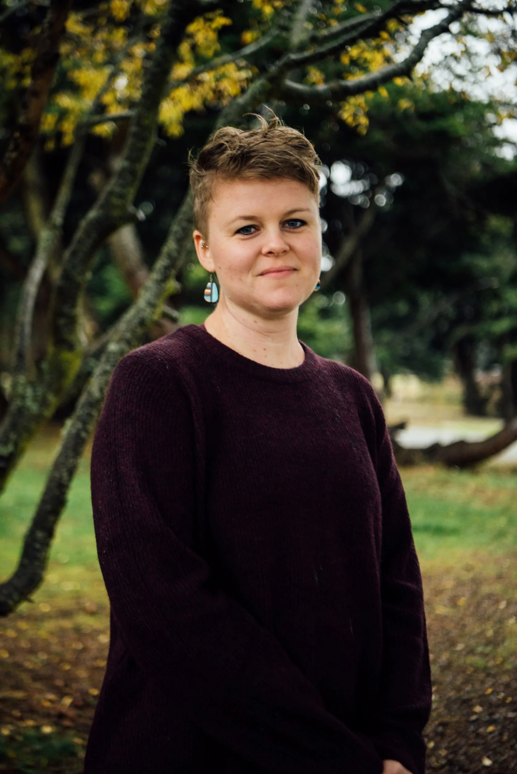A young woman with short hair standing outdoors in front of trees, wearing a dark sweater and colorful earrings, looking at the camera with a slight smile.