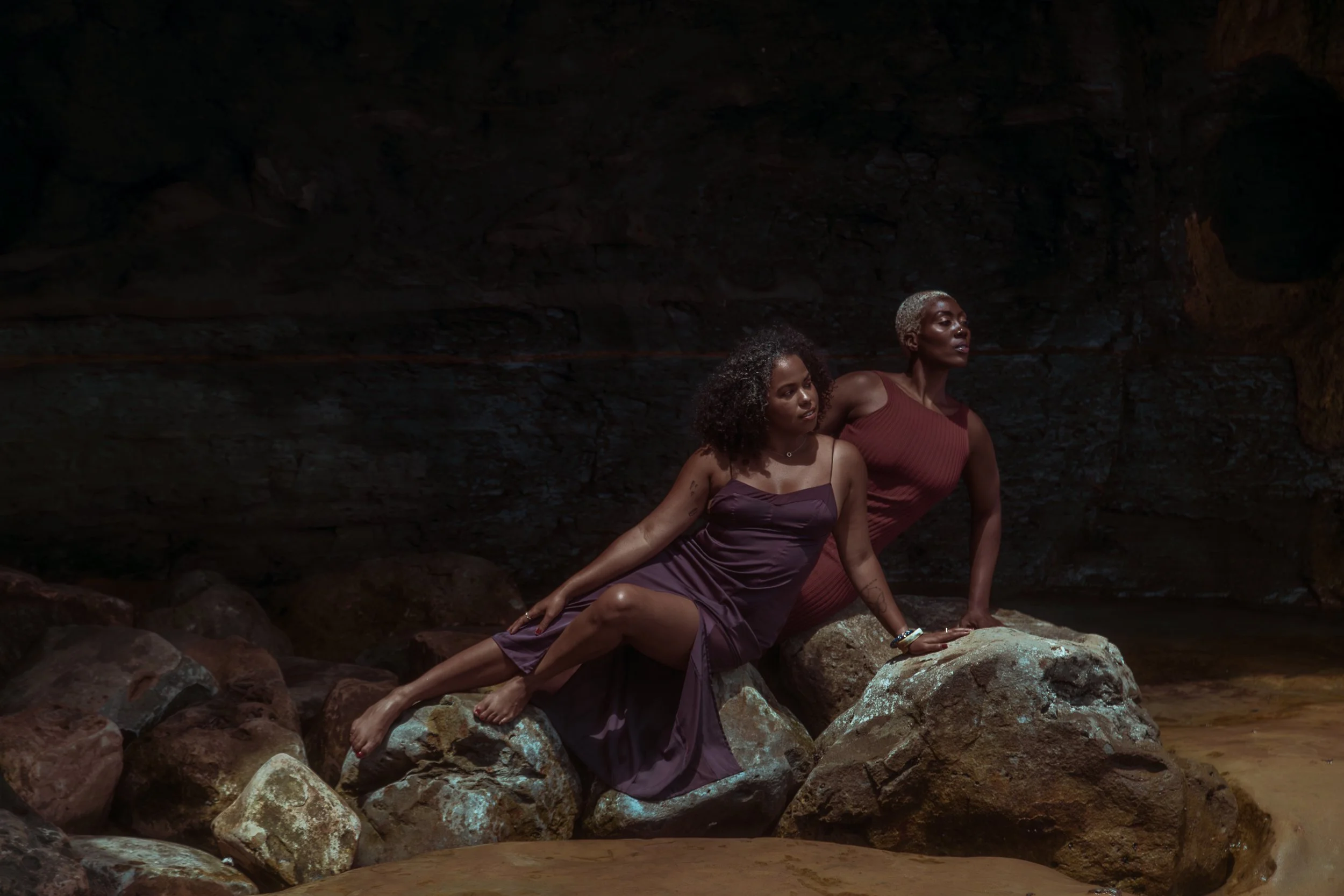 Two women in jewel tone clothing sitting on a rock inside of a cove.