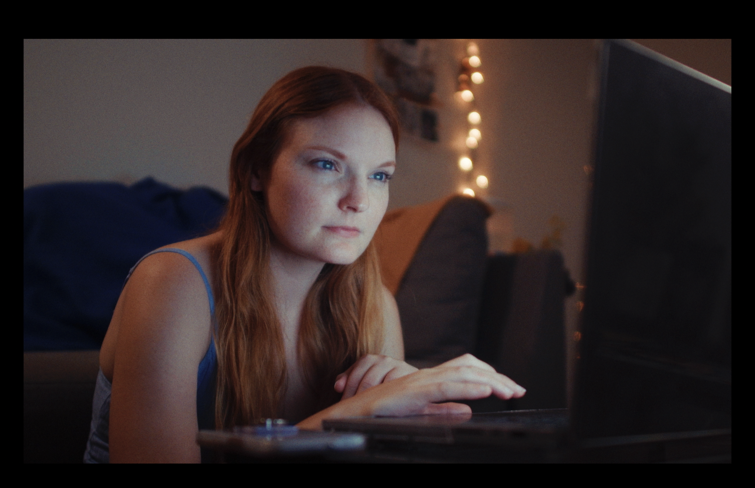 A young woman with red hair and blue eyes looks attentively at a computer screen in a cozy, softly lit room decorated with string lights and personal photos.