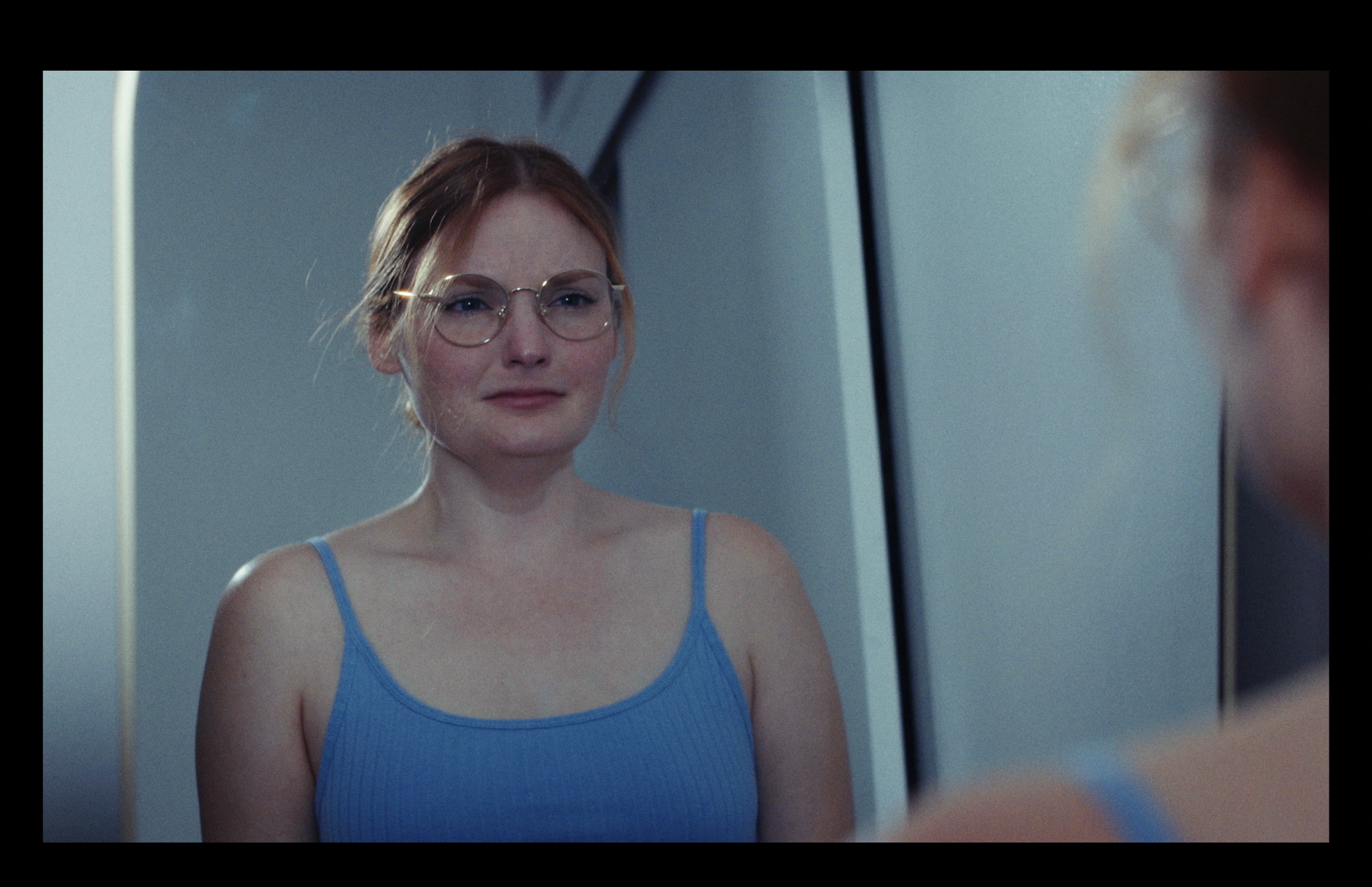 A woman with red hair, glasses, and a blue spaghetti strap top looking at herself in a mirror.