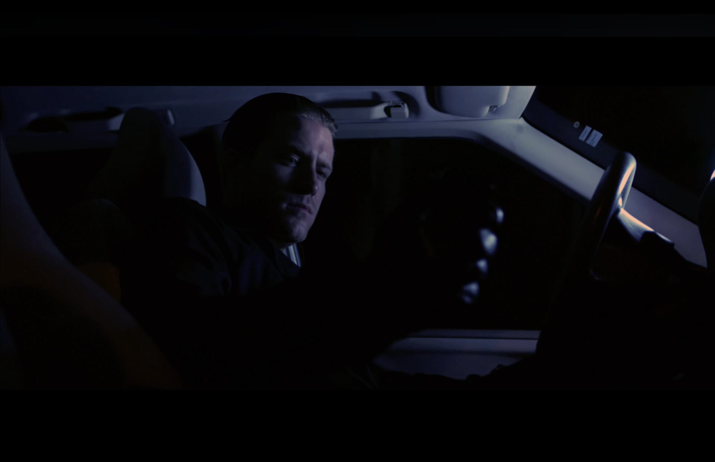 A man sitting in the driver's seat of a car at night, looking toward the camera with a serious expression.