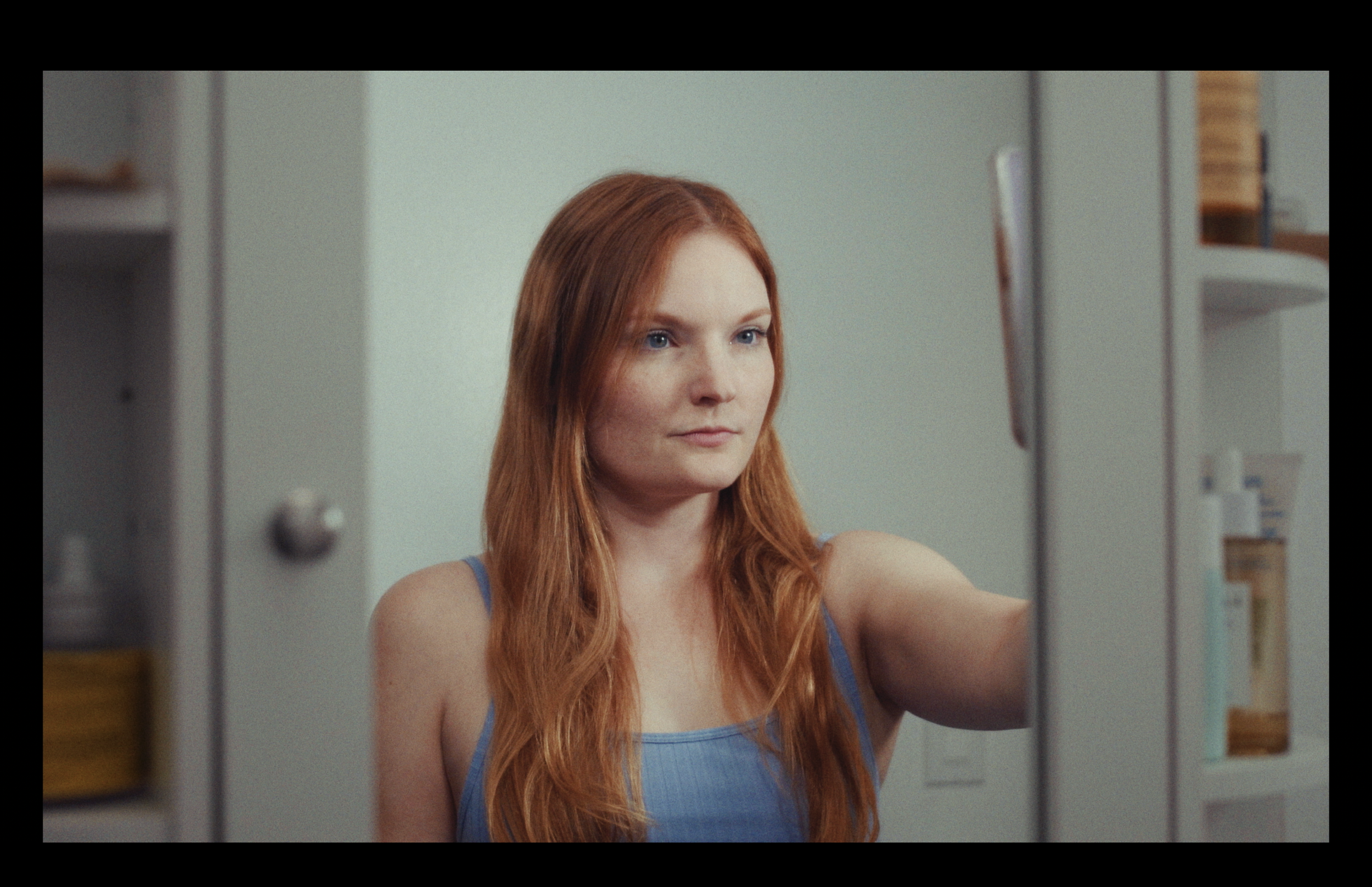 A woman with long red hair and blue eyes, wearing a blue tank top, taking a selfie in the bathroom mirror.
