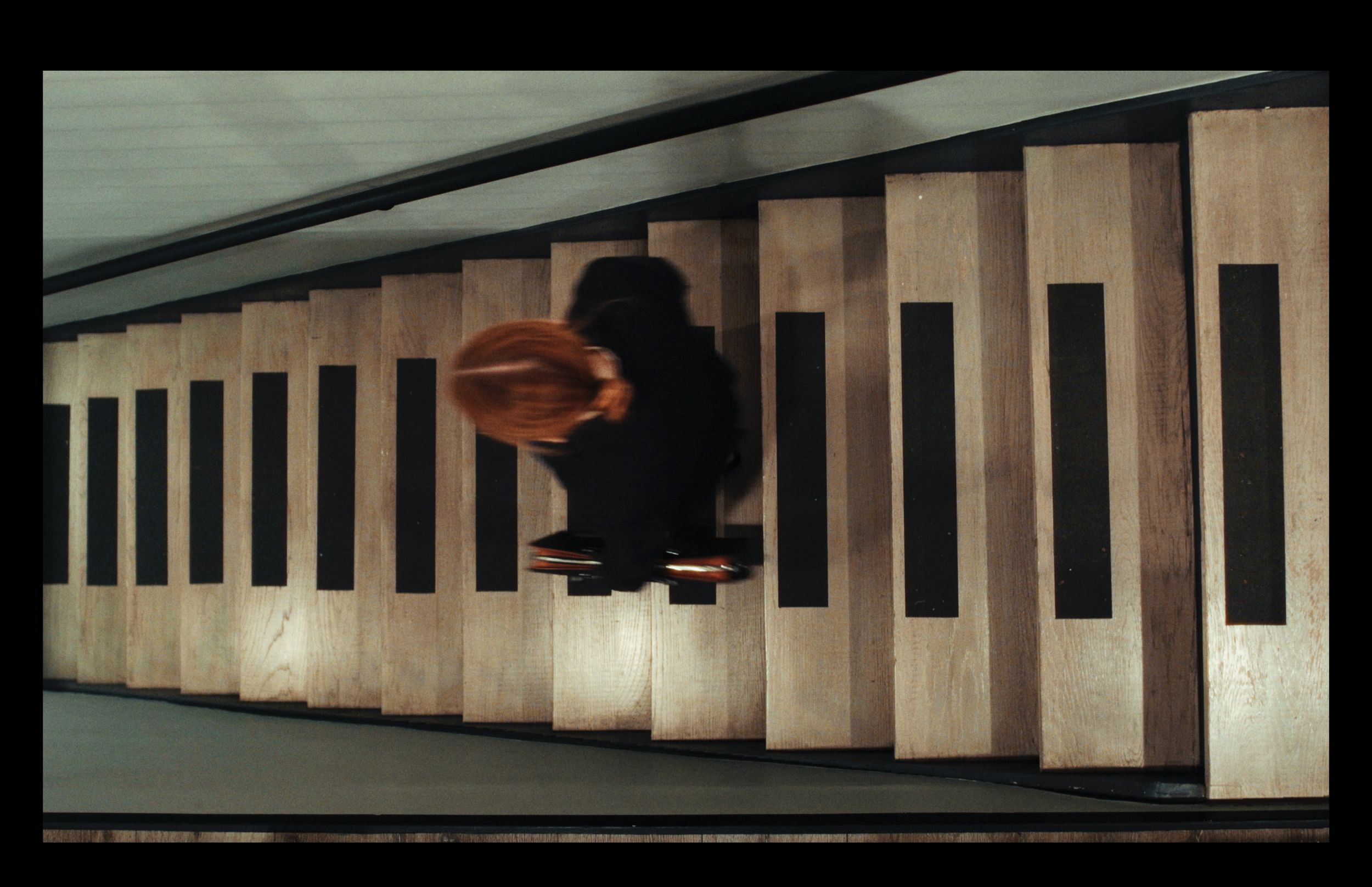 Person with black hair and wearing black clothing climbing down a staircase with wooden steps and black non-slip strips.