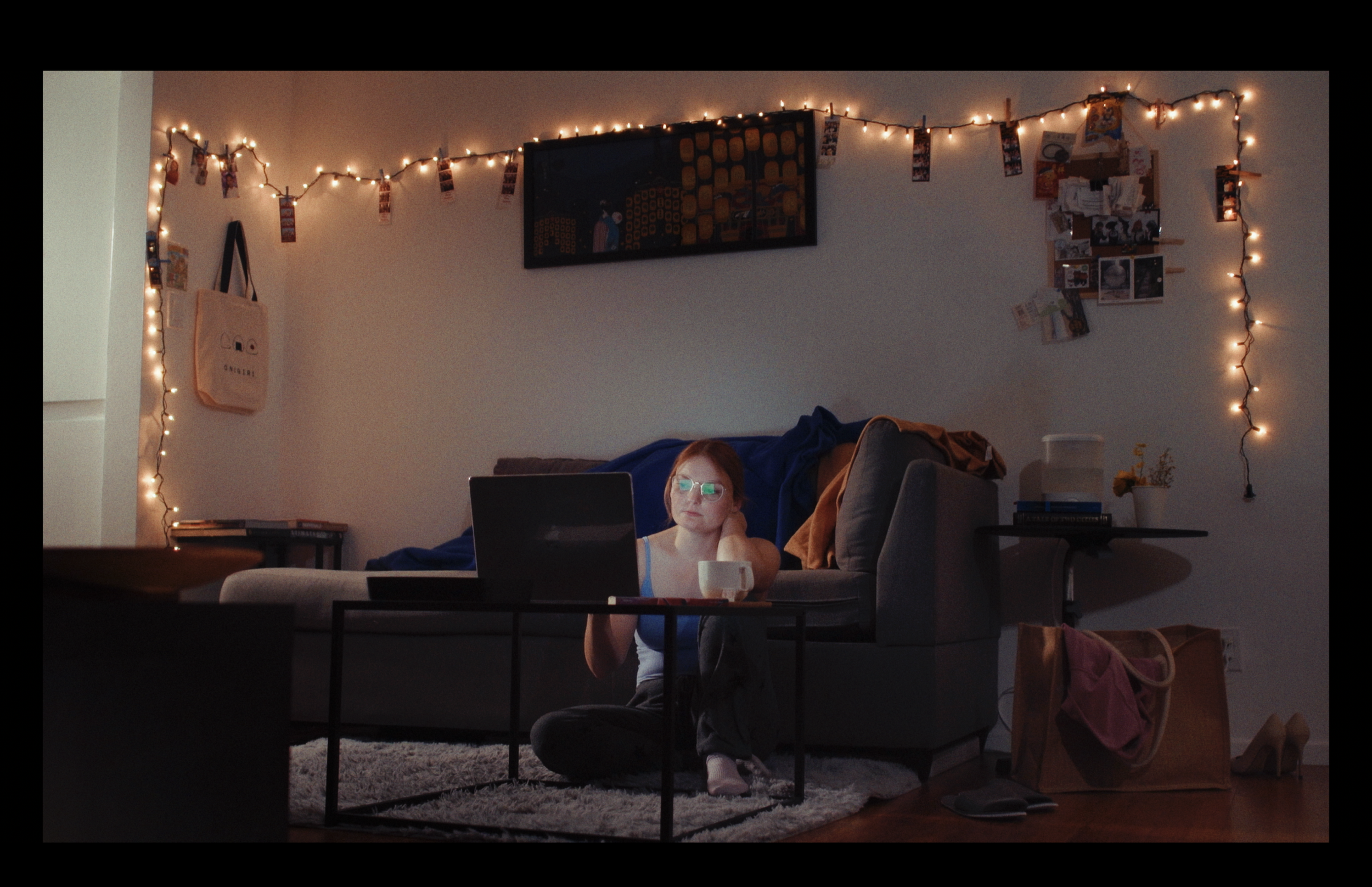 A woman with glasses sits at a table in a dimly lit living room, working on her laptop. The room features string lights, a wall decorated with photos, and various items on shelves and furniture.