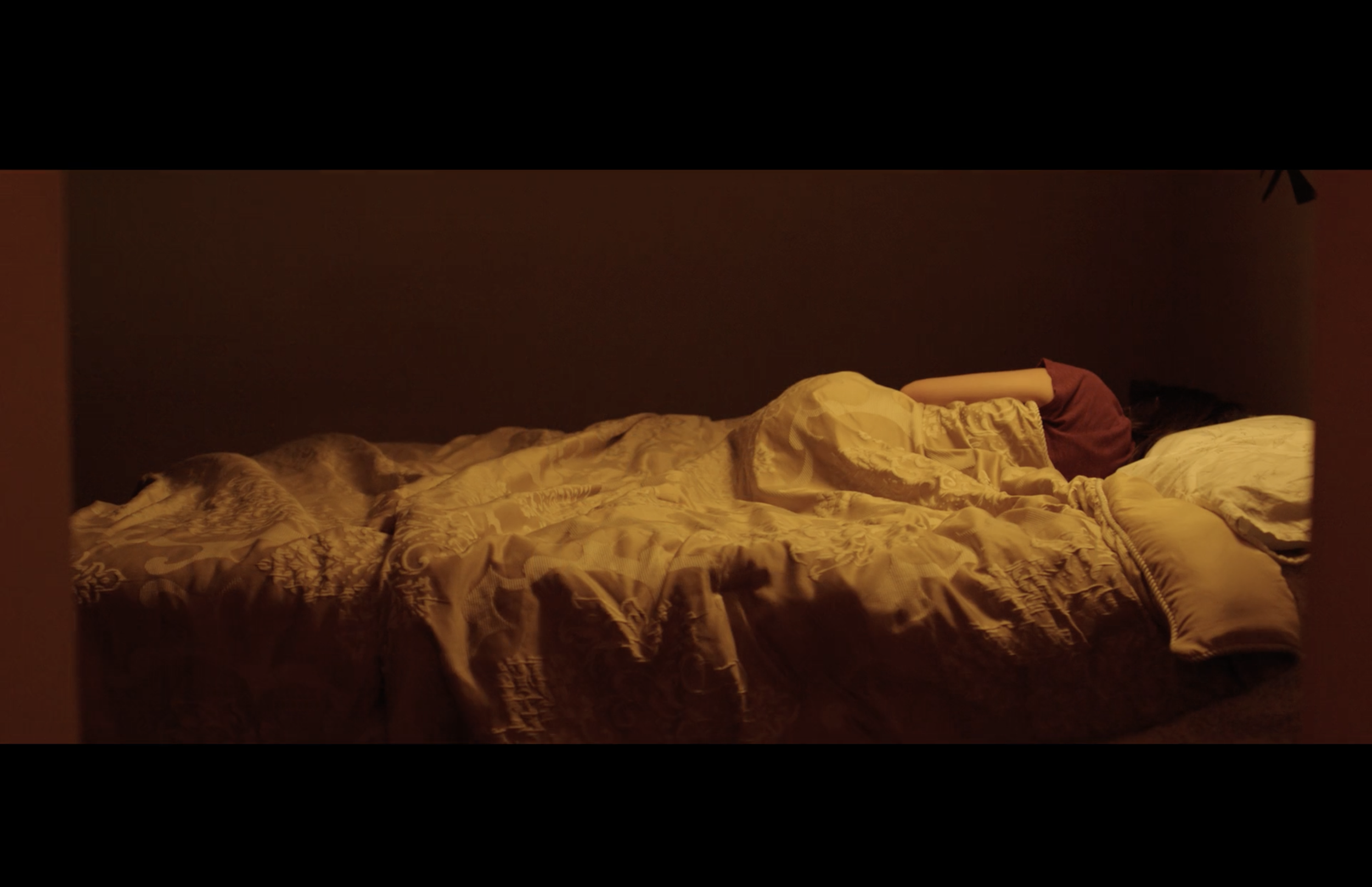 A bed with rumpled yellow patterned sheets and a maroon pillow, in a dimly lit room with a dark wall in the background.