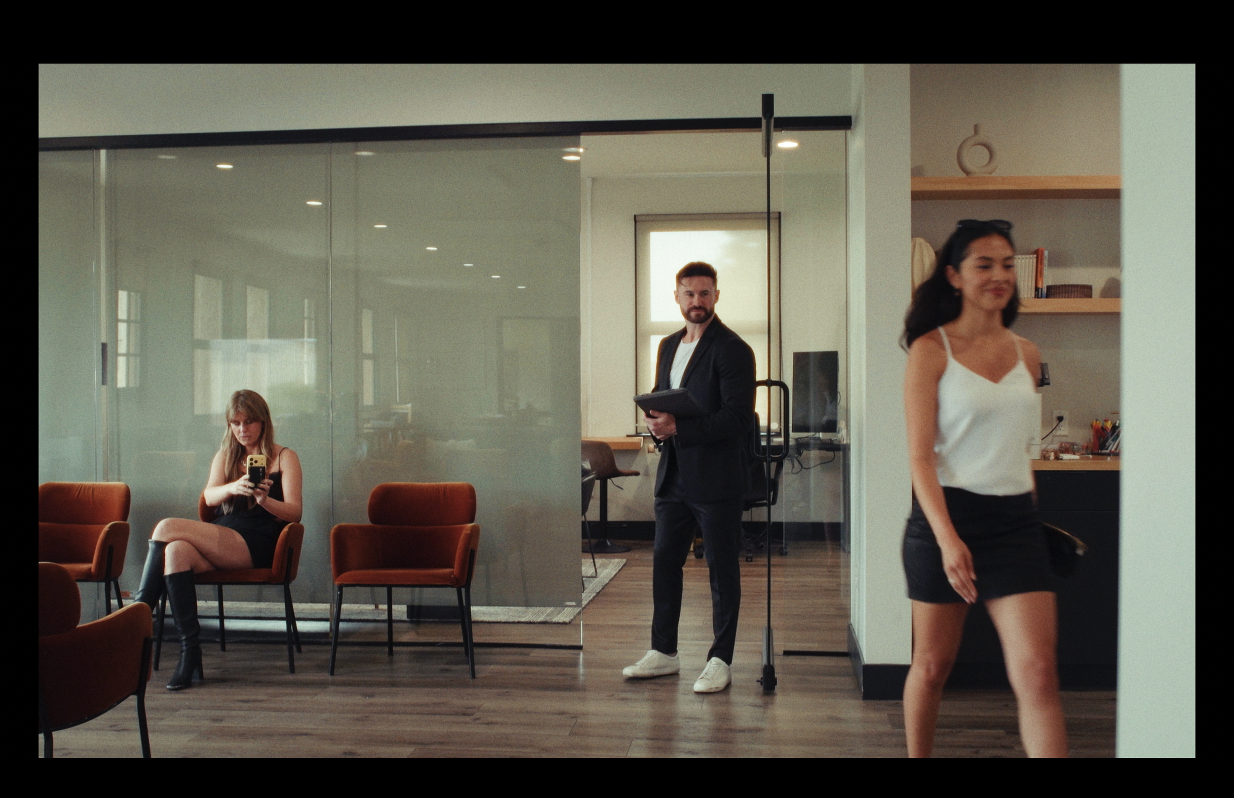 People in a modern office environment, with a woman sitting and looking at her phone, a man standing with a tablet, and a woman walking in front of shelves.