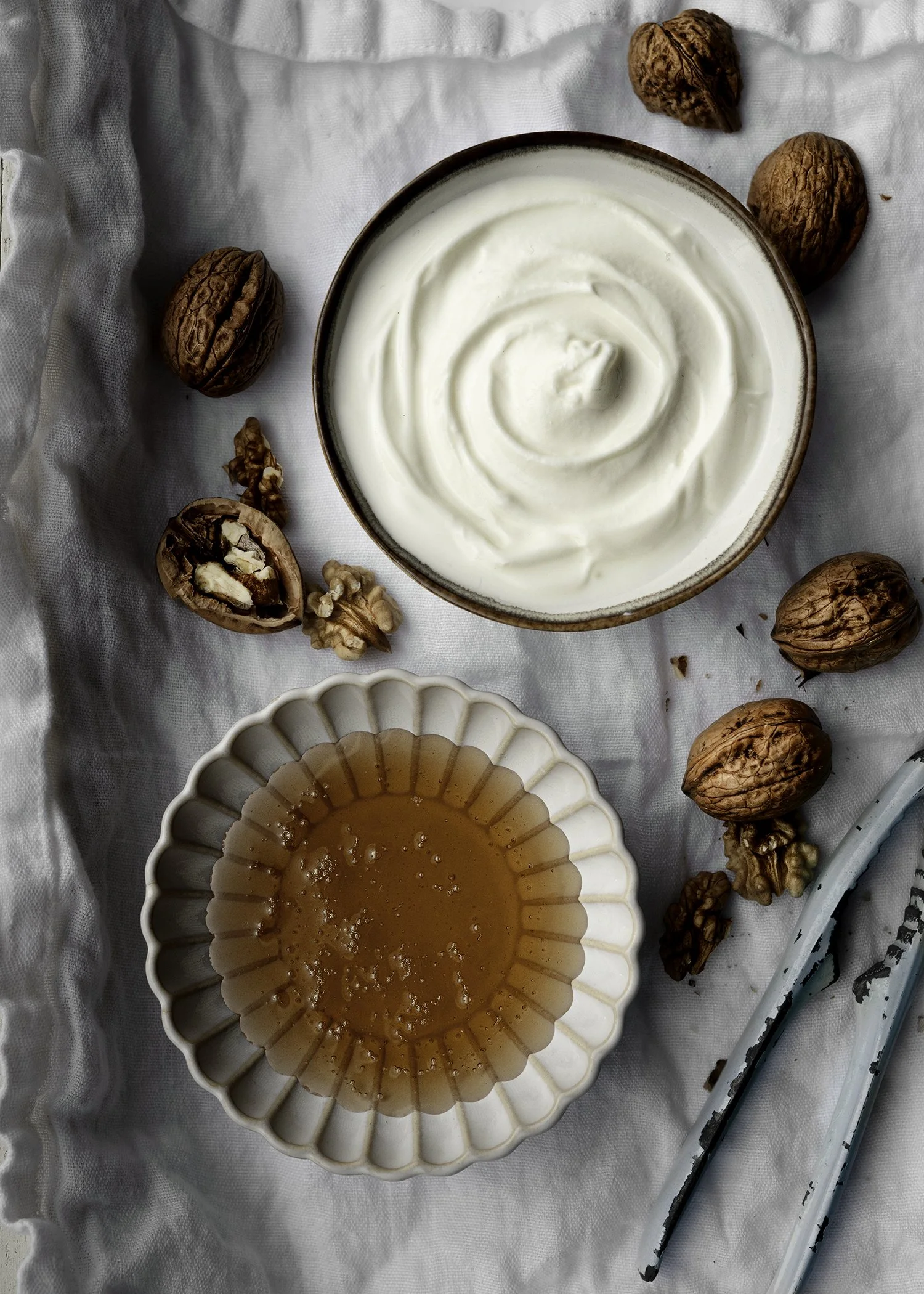 A bowl of creamy white yogurt next to a bowl of honey with two chopsticks on a white cloth, surrounded by whole and halved walnuts.