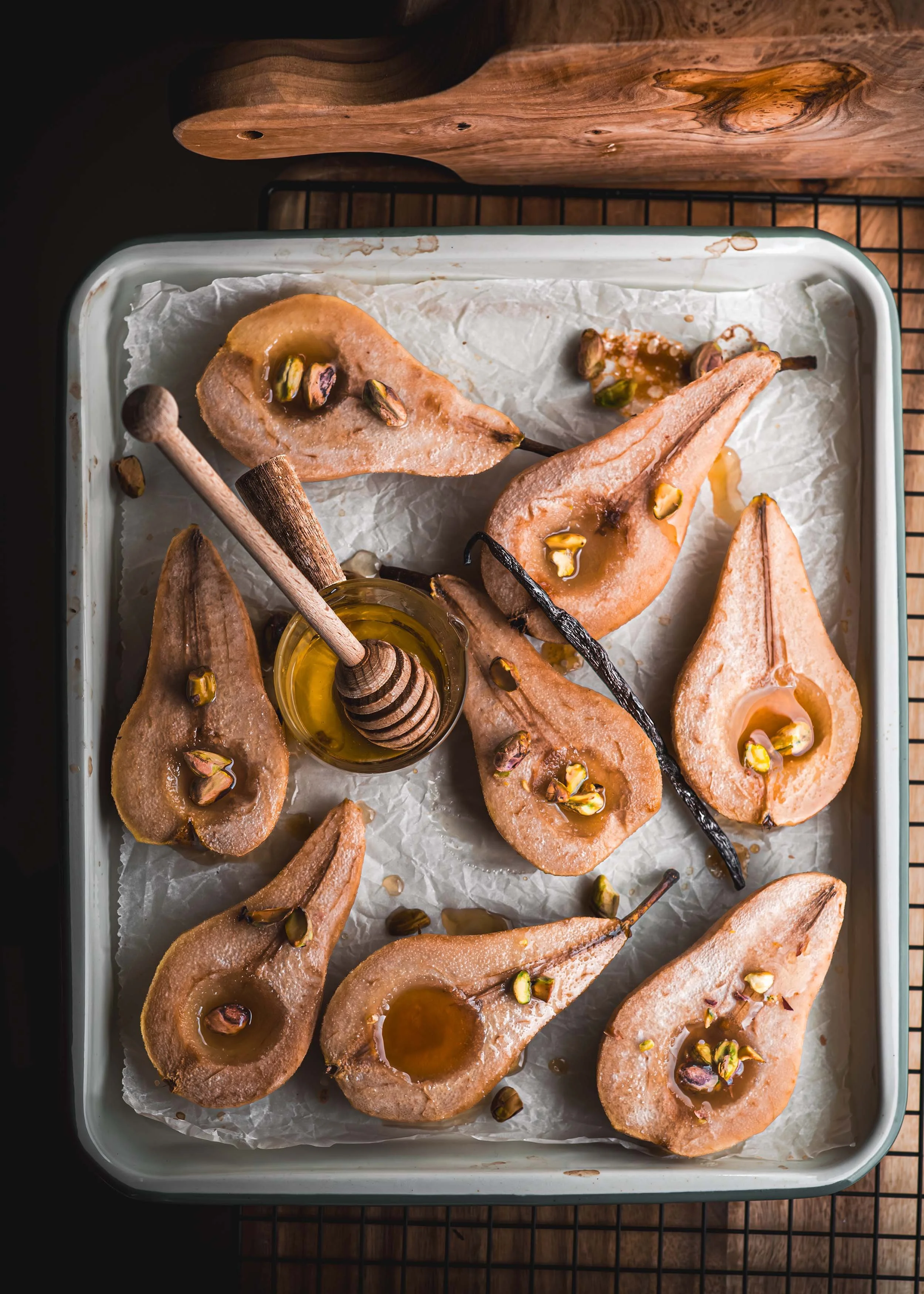 Baked pear halves topped with honey and chopped pistachios, arranged on a white tray lined with parchment paper, with a small jar of honey and a honey dipper.