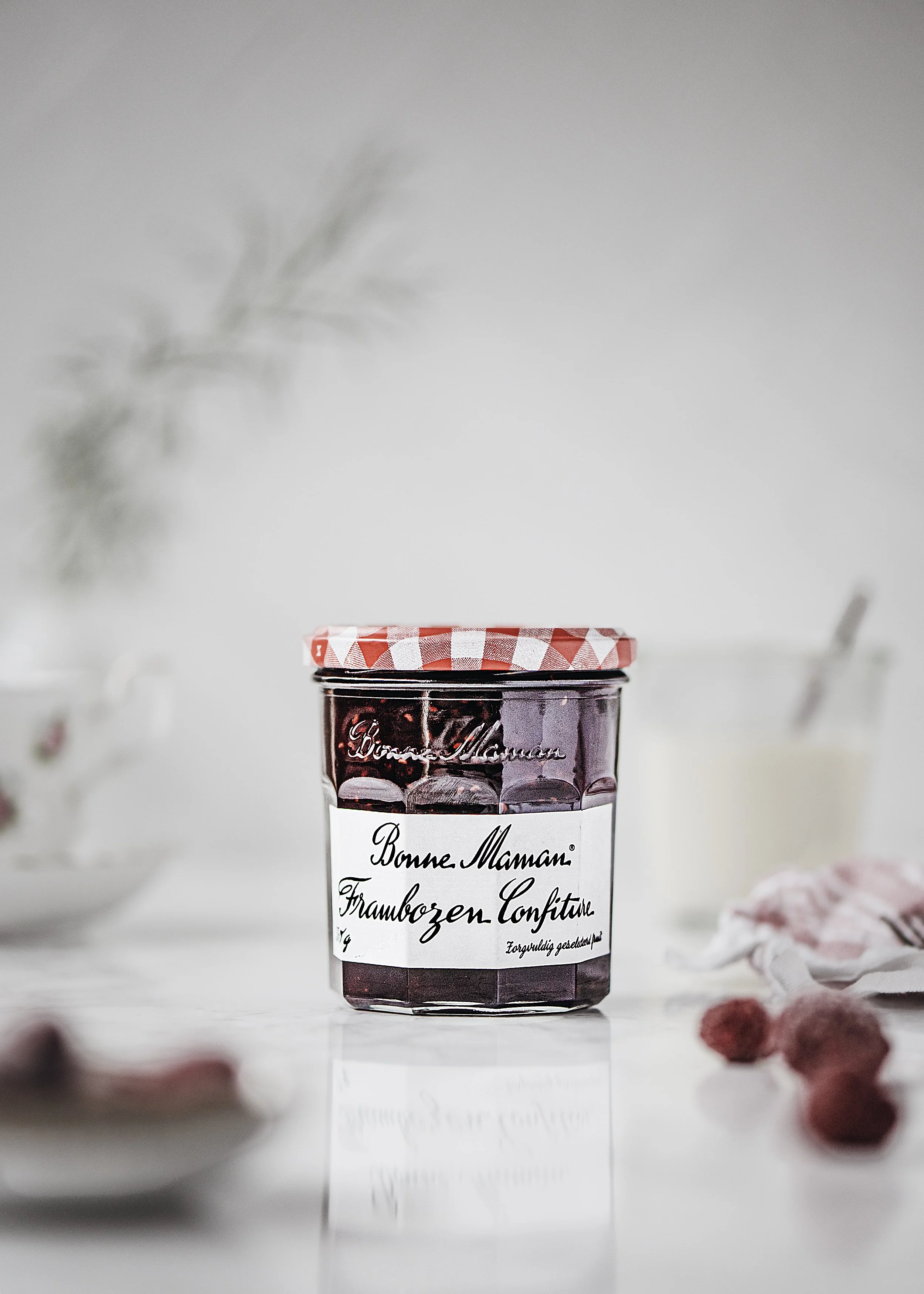 Glass jar of Bonne Maman strawberry freezer jam with red and white checkered lid, placed on a white surface with blurred raspberries and dried flowers in the background.