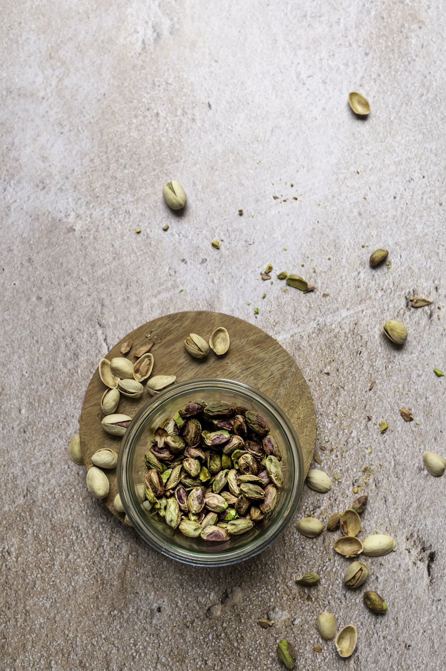 A glass bowl filled with shelled pistachios on a round wooden board, with scattered pistachio shells and pistachos around the bowl on a textured surface.