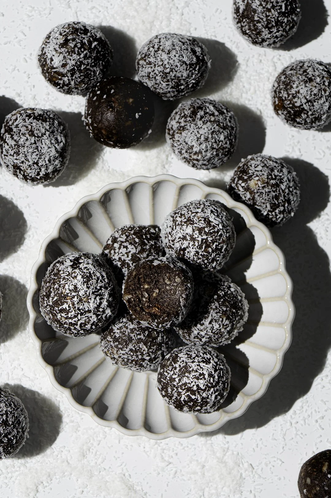 Chocolate energy bites topped with shredded coconut, some on a white dish and others scattered on a white surface.
