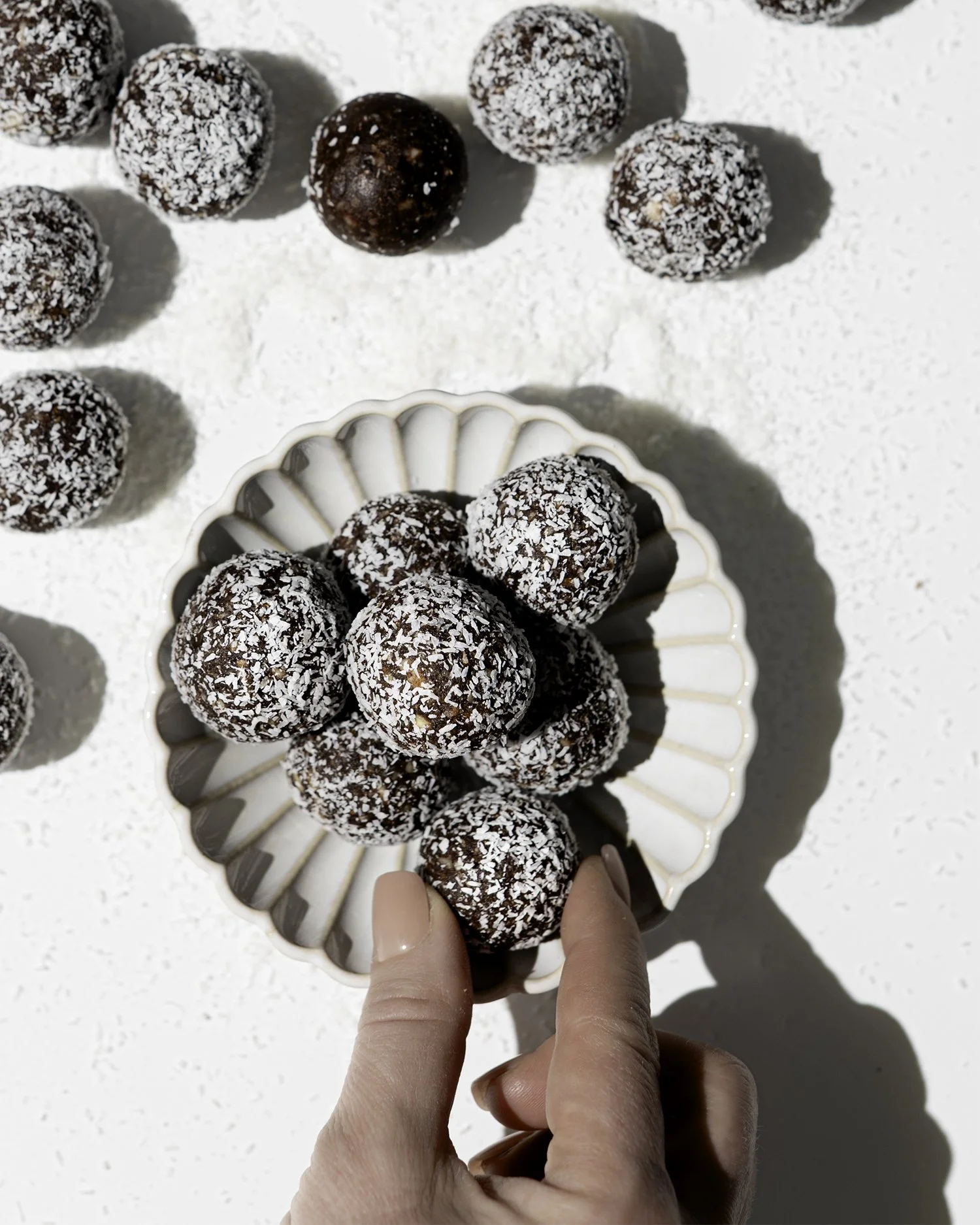 Chocolate energy balls covered with shredded coconut on a white plate, with one being held by two fingers.