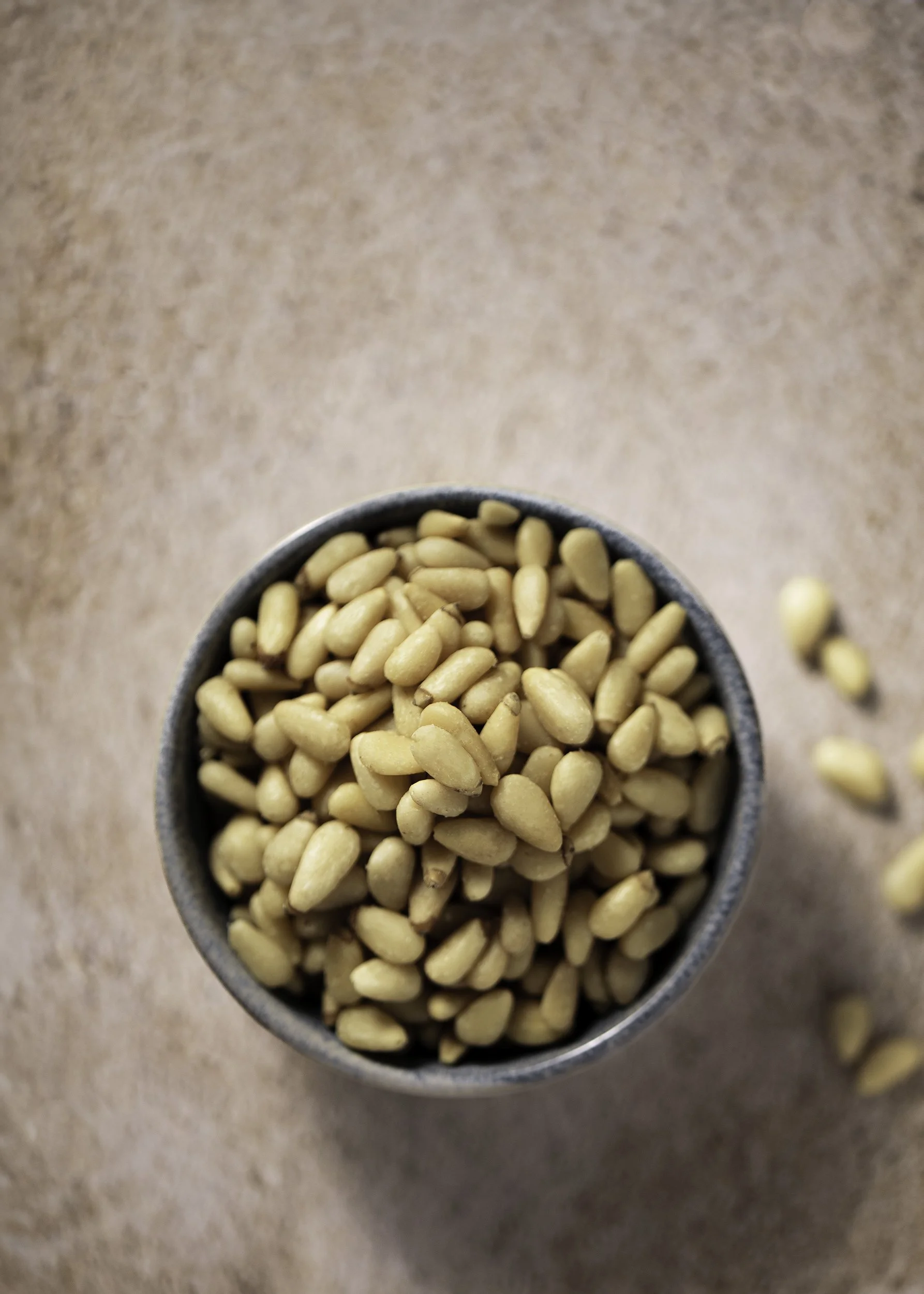 A top view of a small gray bowl filled with pine nuts on a brown textured surface.