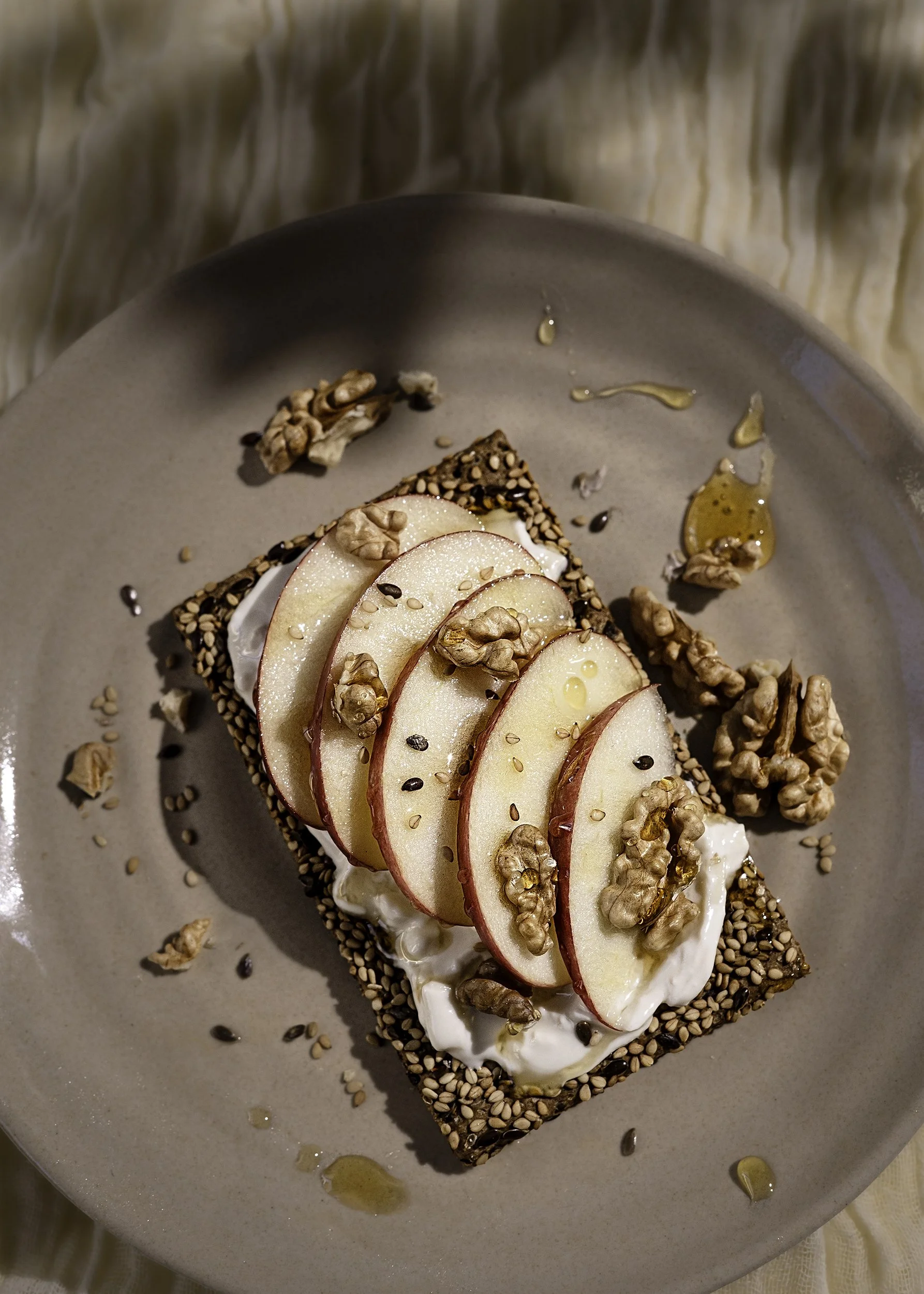 Sliced pears on top of a piece of bread with spread, walnuts, sesame seeds, and honey, served on a beige plate.