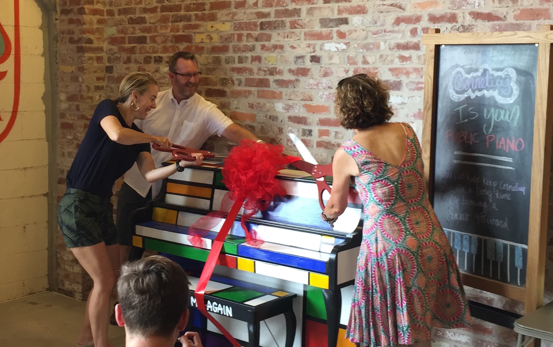 Beth hodges (piano donor) and theresa dean (piano artists) cutting the ribbon as "cornelius" is introduced to ponce city market.