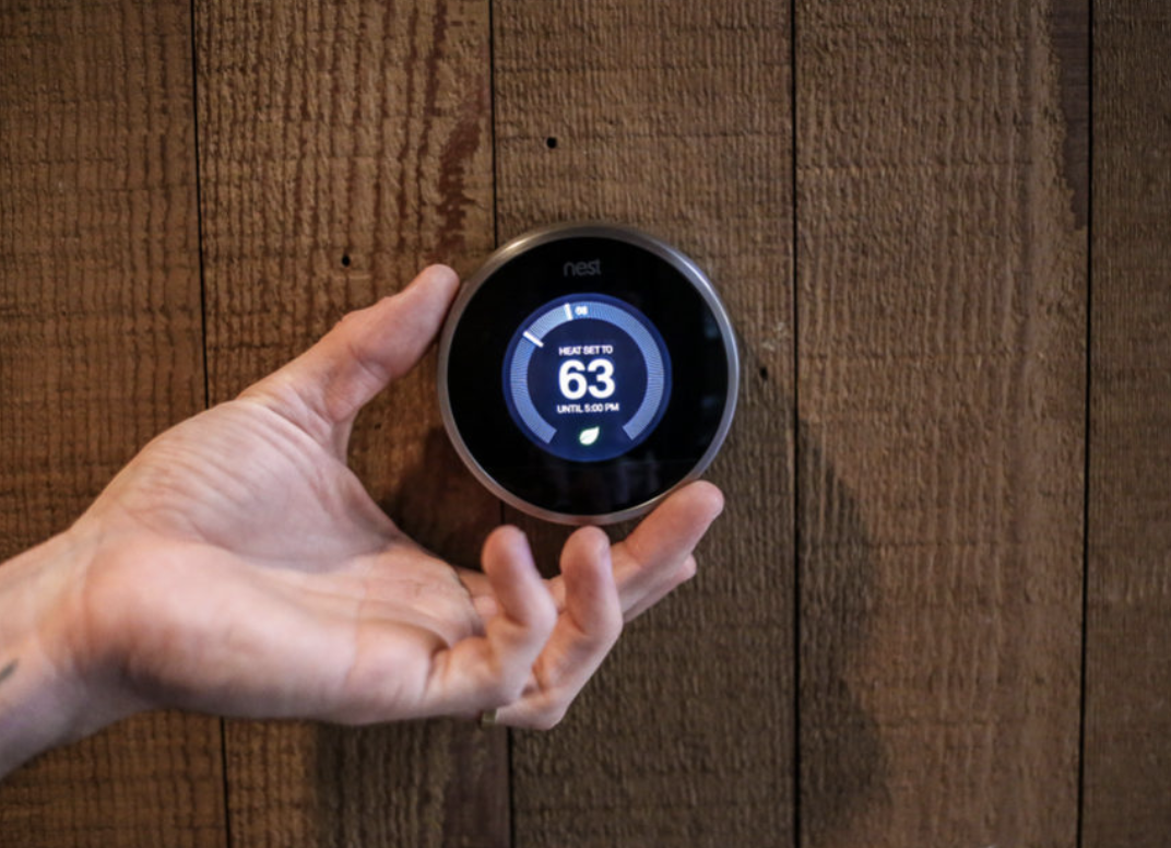 Hand adjusting a Nest smart thermostat on a wooden wall.