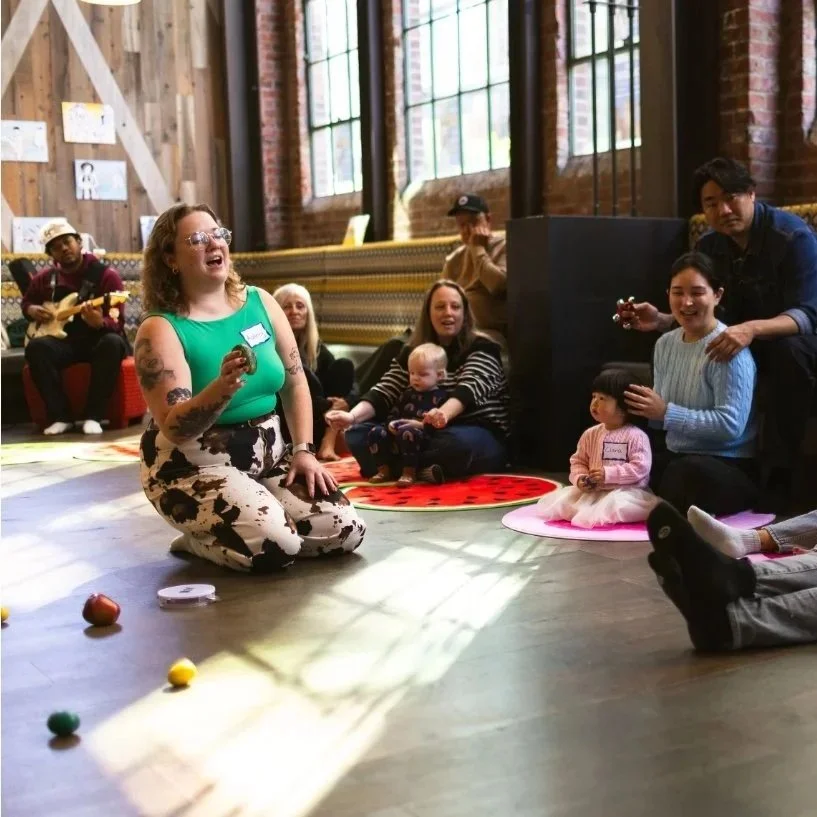 Teacher in a green top holding an egg shaker, singing in front of babies, toddlers, and their parents. Photo by Parker Miles Blohm