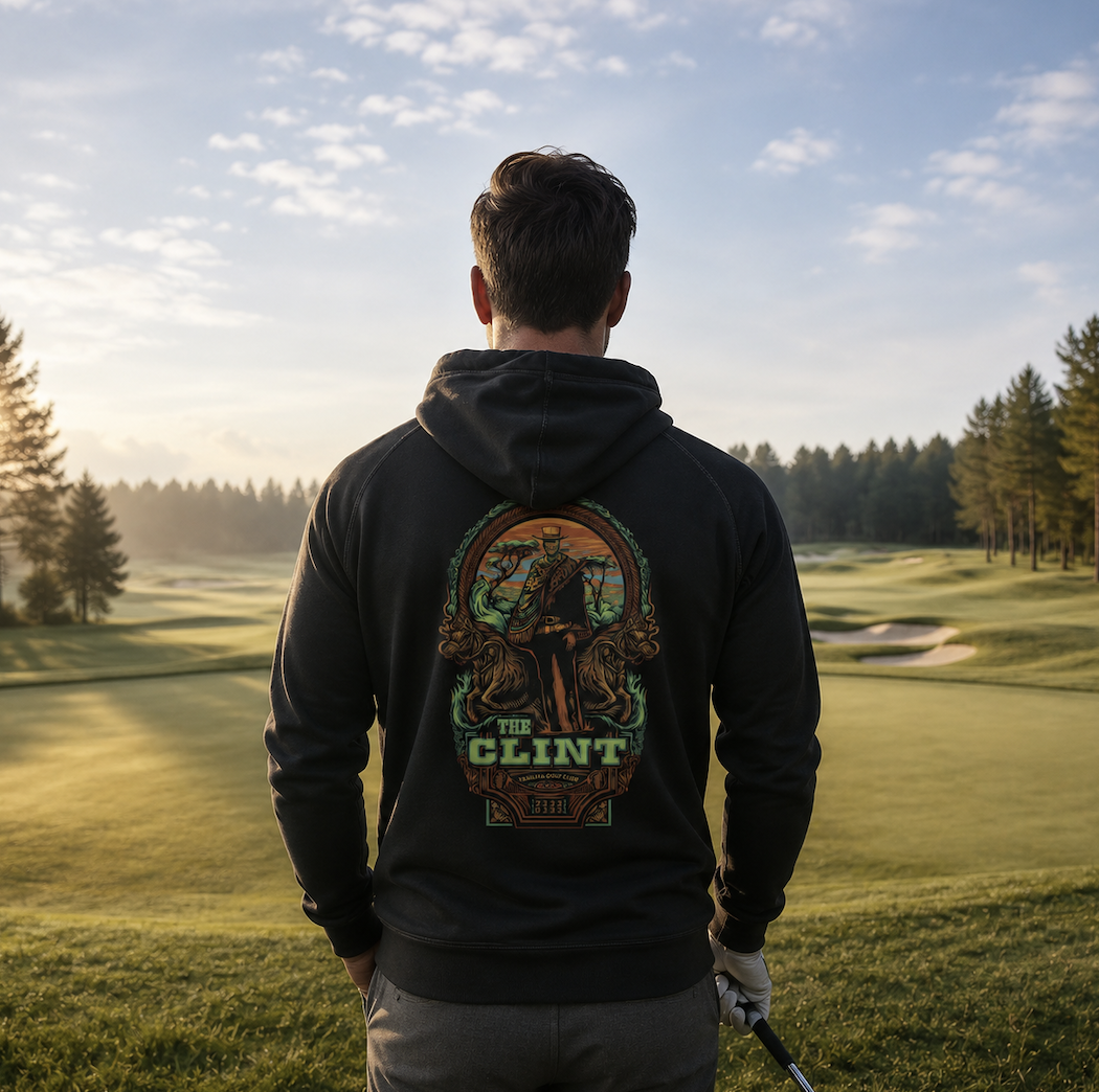A man in a black hoodie with a colorful graphic on the back, holding a golf club, standing on a golf course during the day with trees and sand traps.
