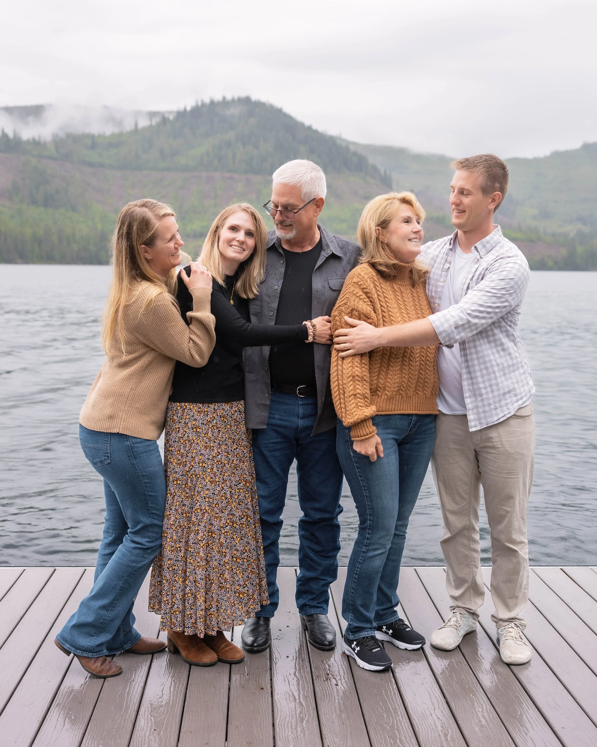Family standing together on a dock by a lake with misty mountain views during an outdoor family photo session in North Idaho.
