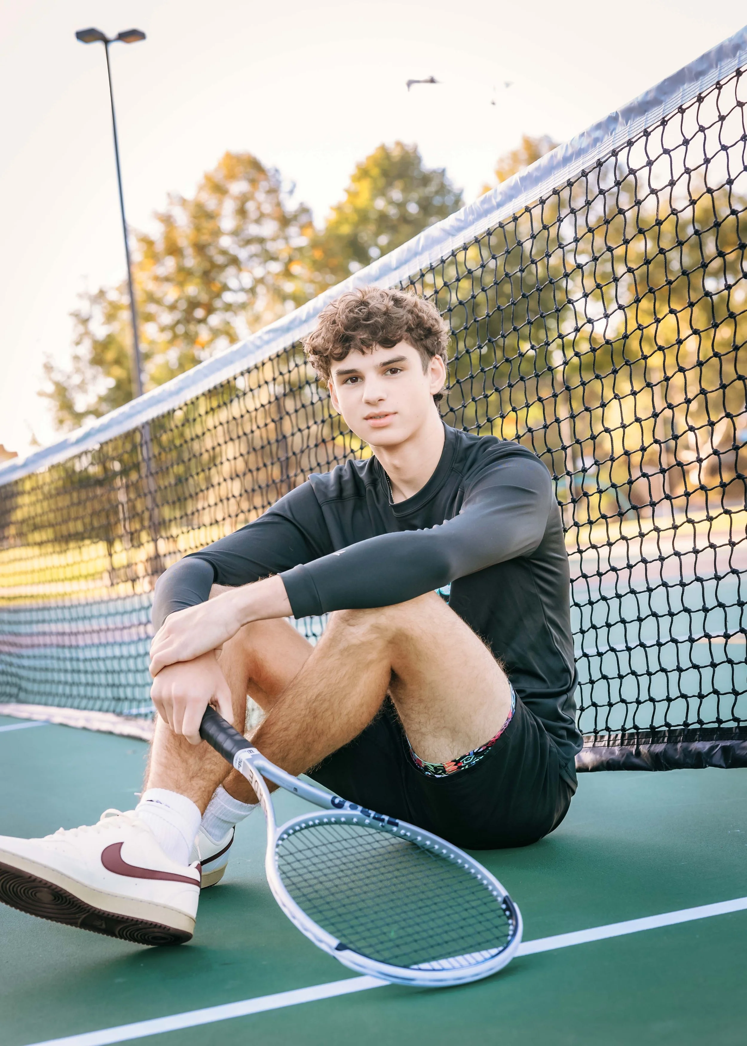 High school senior sitting on a tennis court holding a tennis racket during a personalized athletic senior photo session in North Idaho.