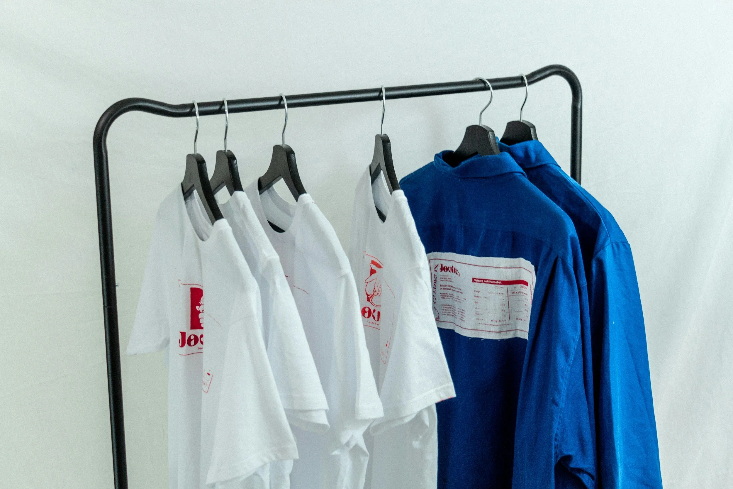 A clothing rack holding four white t-shirts and two blue shirts on black hangers against a plain white wall.