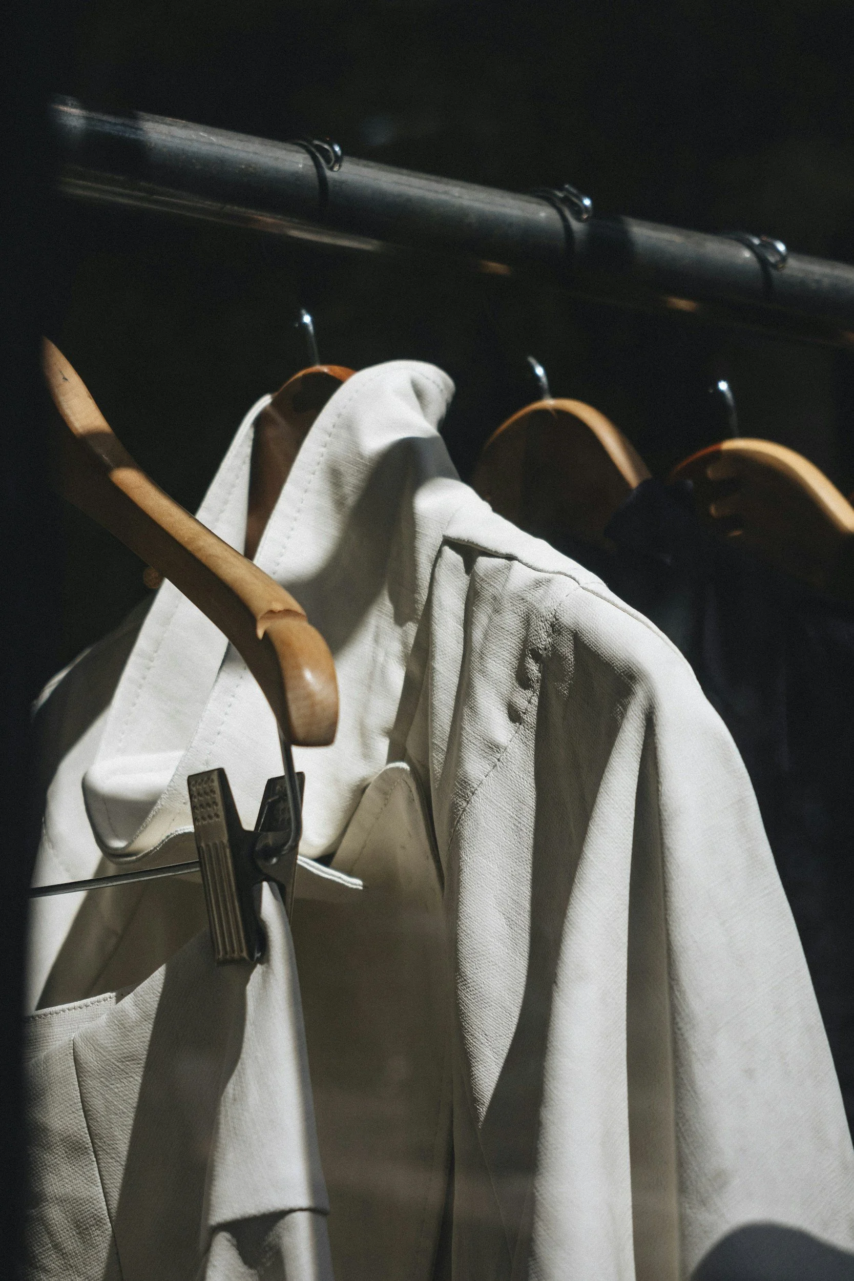 White jacket hanging on a wooden hanger, with other clothing items on hangers behind it, on a clothing rack.