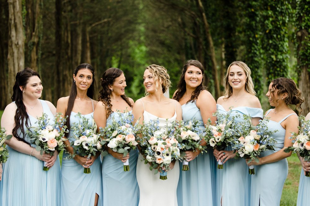  Candid photo of bride with her bridesmaids, in light blue dresses holding white, peach, and blue bouquets 