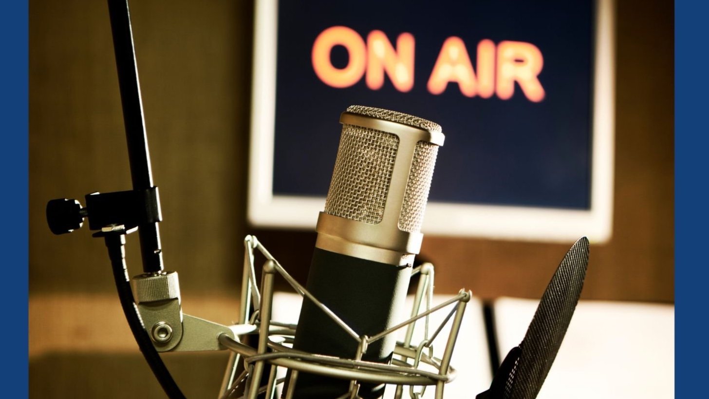 A professional microphone in a recording studio is in front of a screen with a red lit 'ON AIR' sign, indicating live broadcasting.