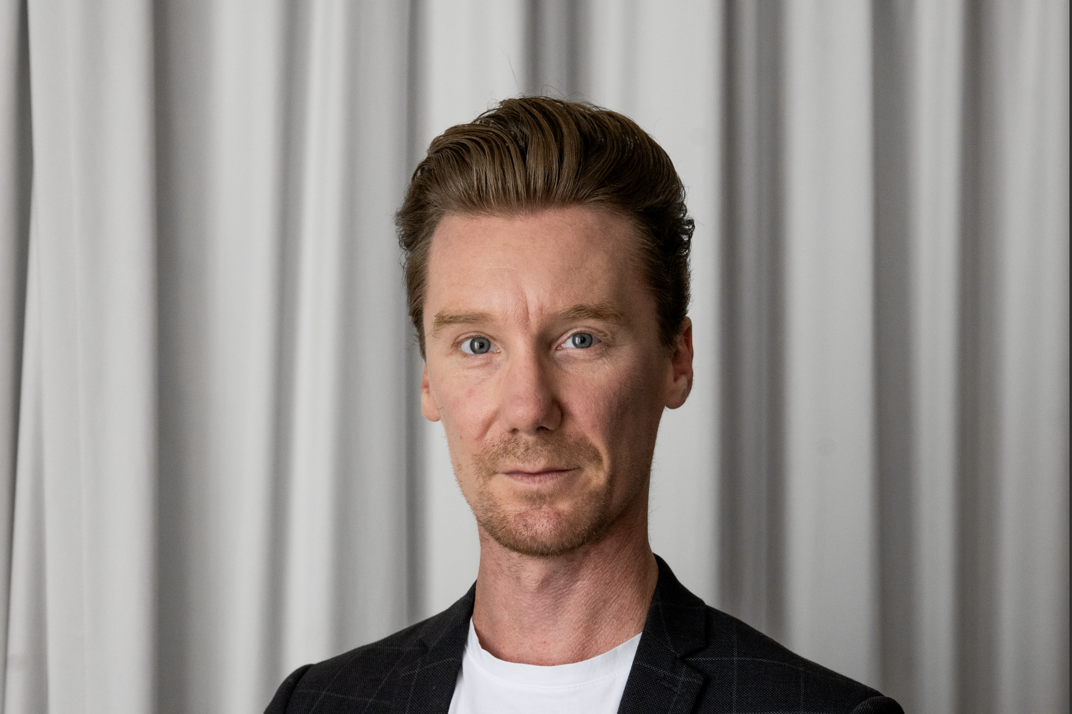 Luke Butler (He/him), is the Vice President of Communications at DPA. He has short auburn hair, blue eyes, short facial hair and is wearing a blue blazer with a white shirt. He has a steely gaze.