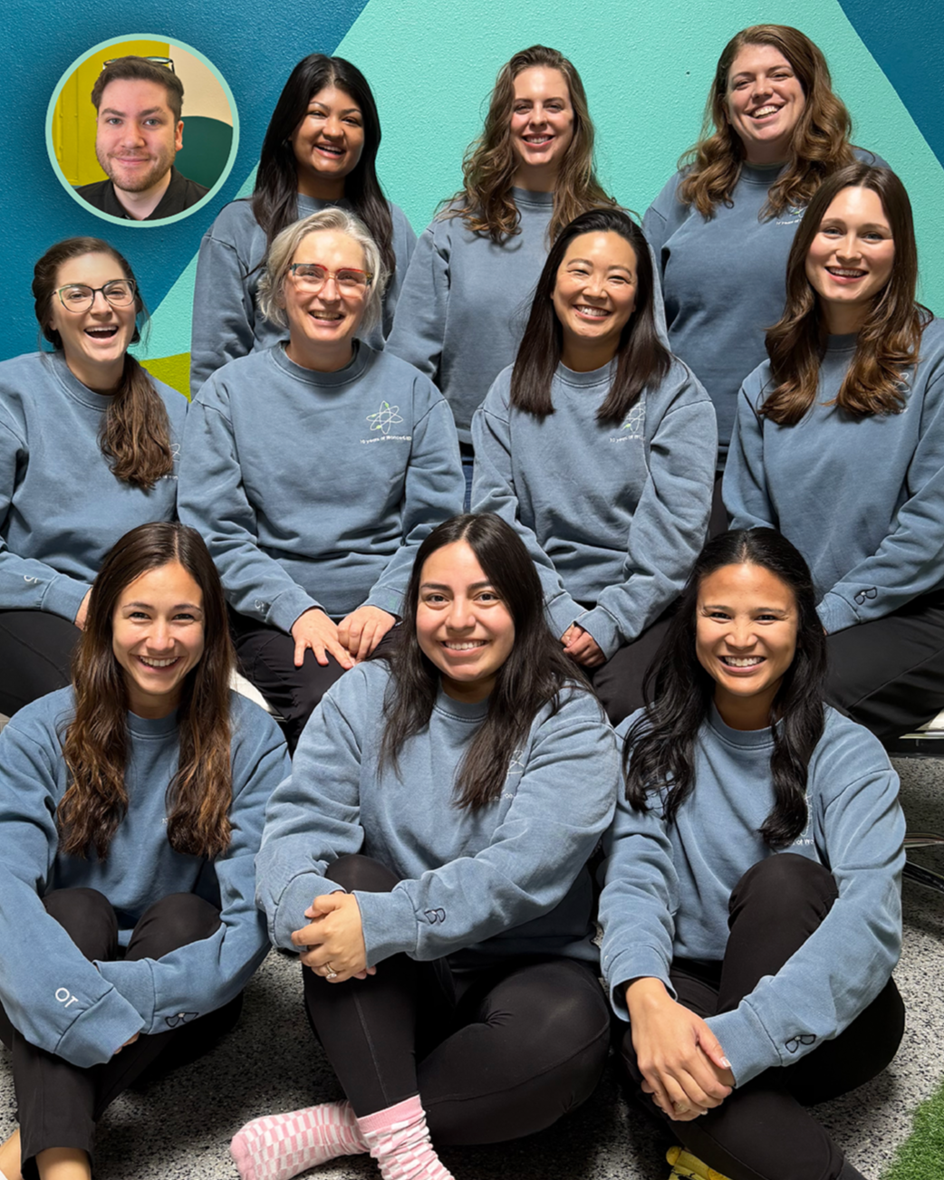 Group of nine women and one man posing for a photo, all wearing matching light blue sweatshirts, in front of a colorful wall with geometric shapes.