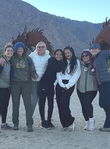 Group of seven women smiling and posing outdoors with mountainous background.