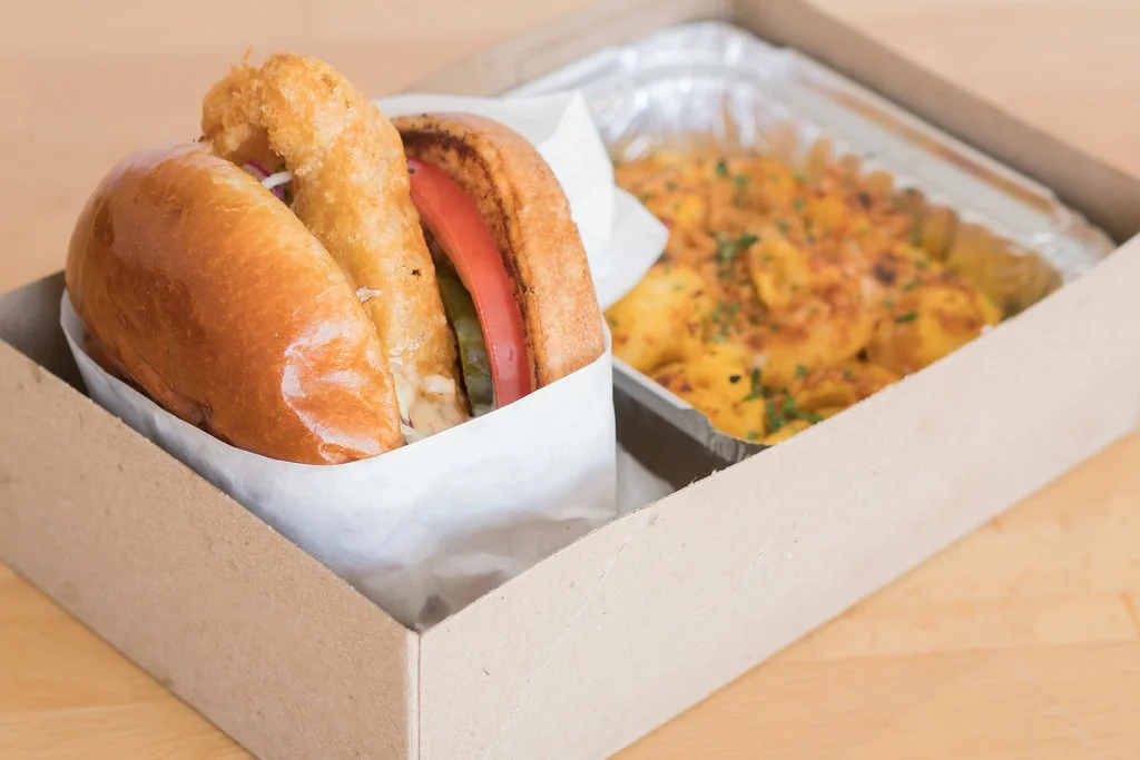 Boxed catering meal with a fried chicken sandwich, tomato slice, and tray of baked macaroni and cheese.