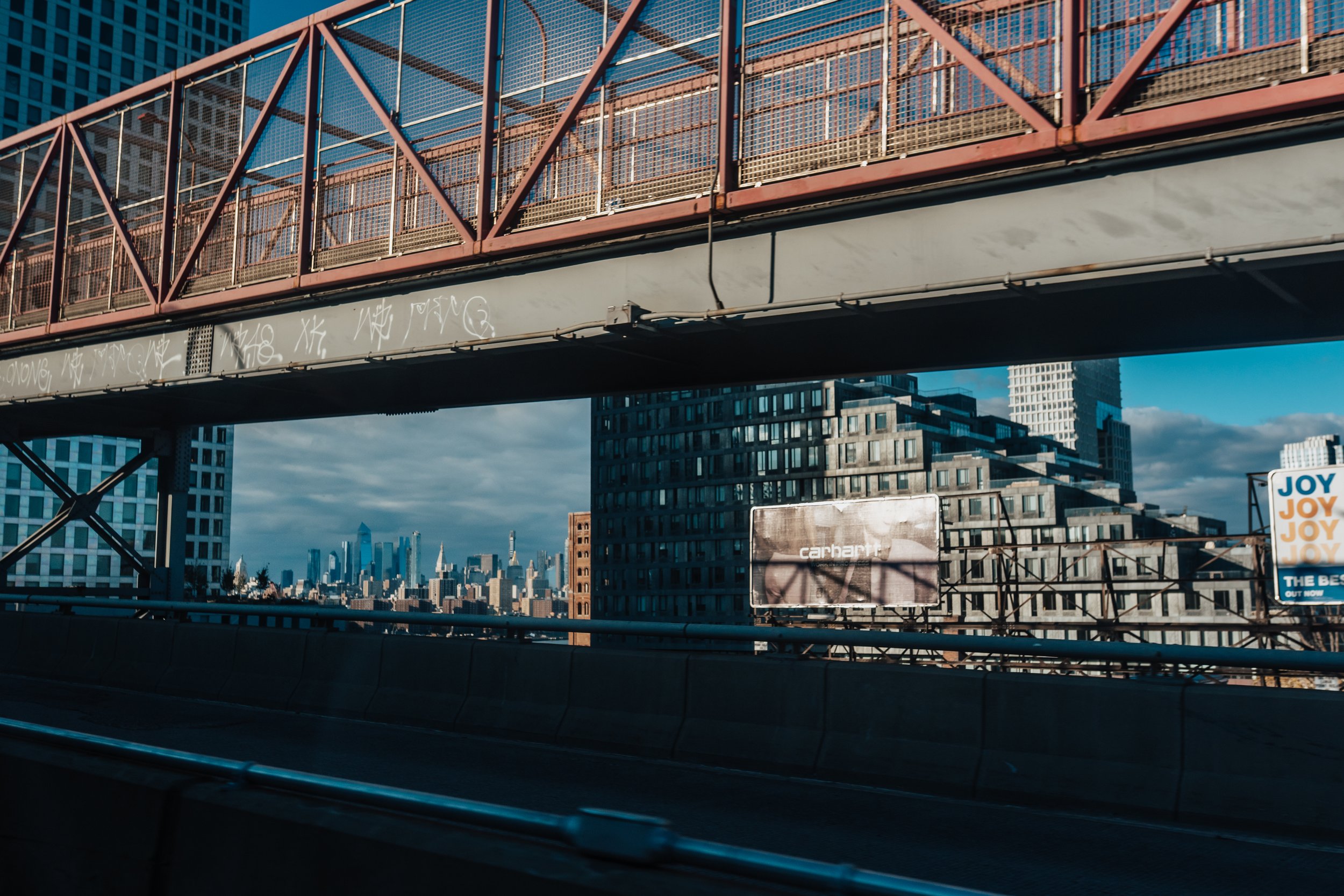 View of a city skyline seen from a highway, with buildings and skyscrapers in the distance, a pedestrian bridge above the highway, and a cloudy sky.