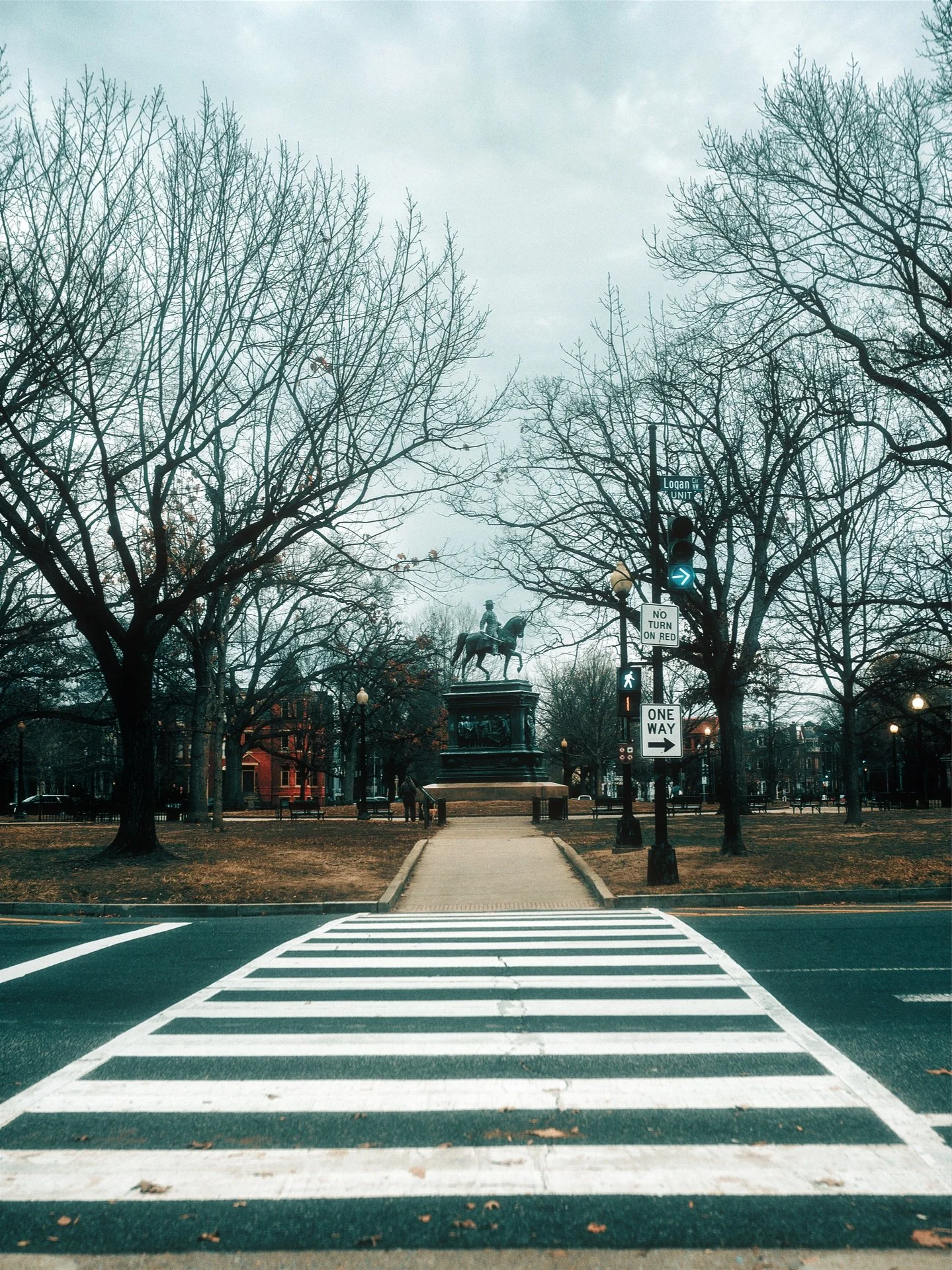 A crosswalk leading to a park with leafless trees, a statue of a horse and rider, and street signs including one for Logan Circle.