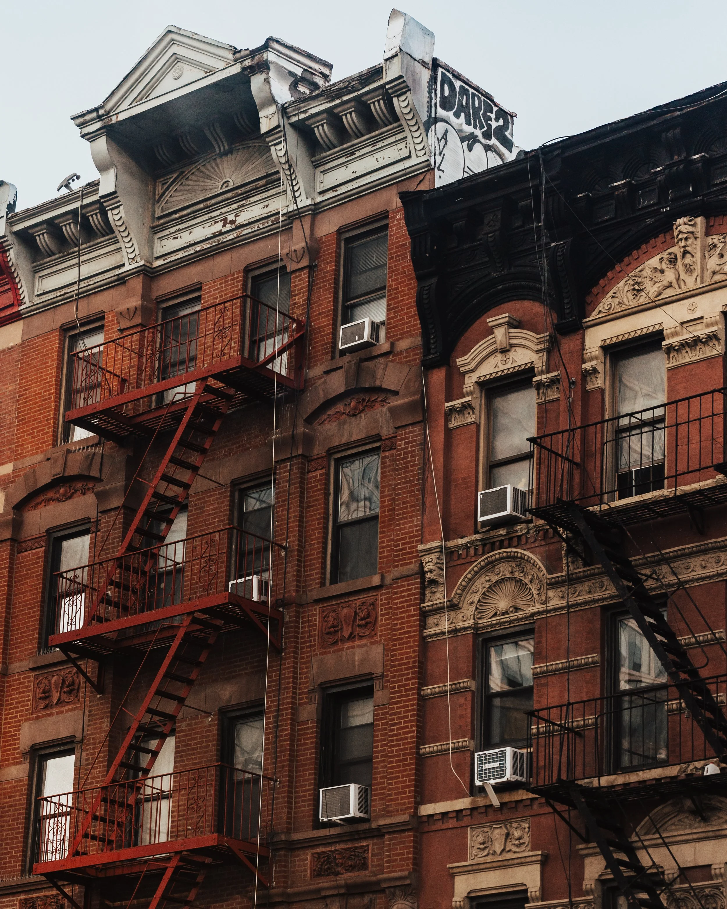 Close-up of a historic brick apartment building with fire escapes, ornate architectural details, and air conditioning units in the windows.