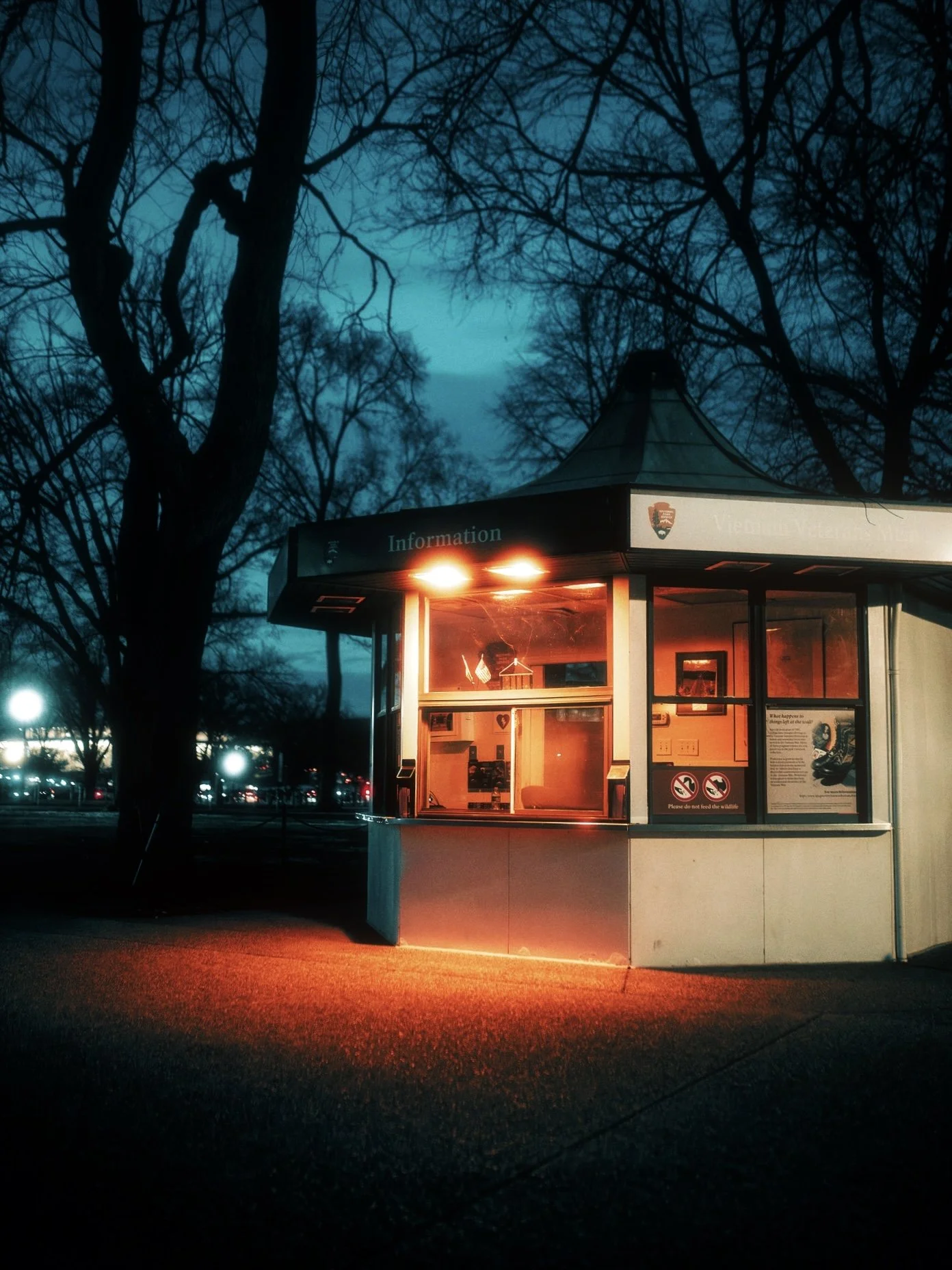 A small, well-lit visitor information kiosk at dusk with no people inside, surrounded by leafless trees and a darkening sky.