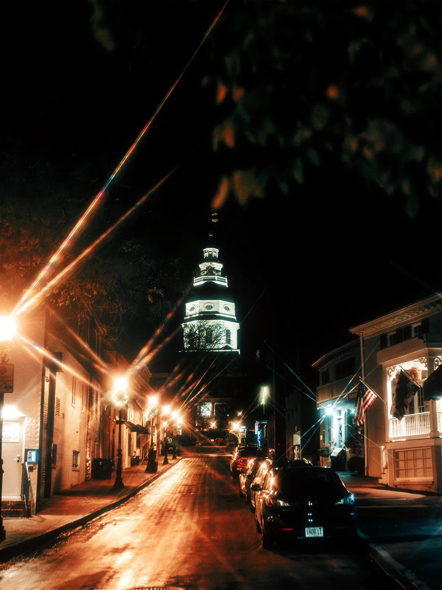 Nighttime city street scene with a lit tower in the background and streetlights illuminating the road and sidewalk.