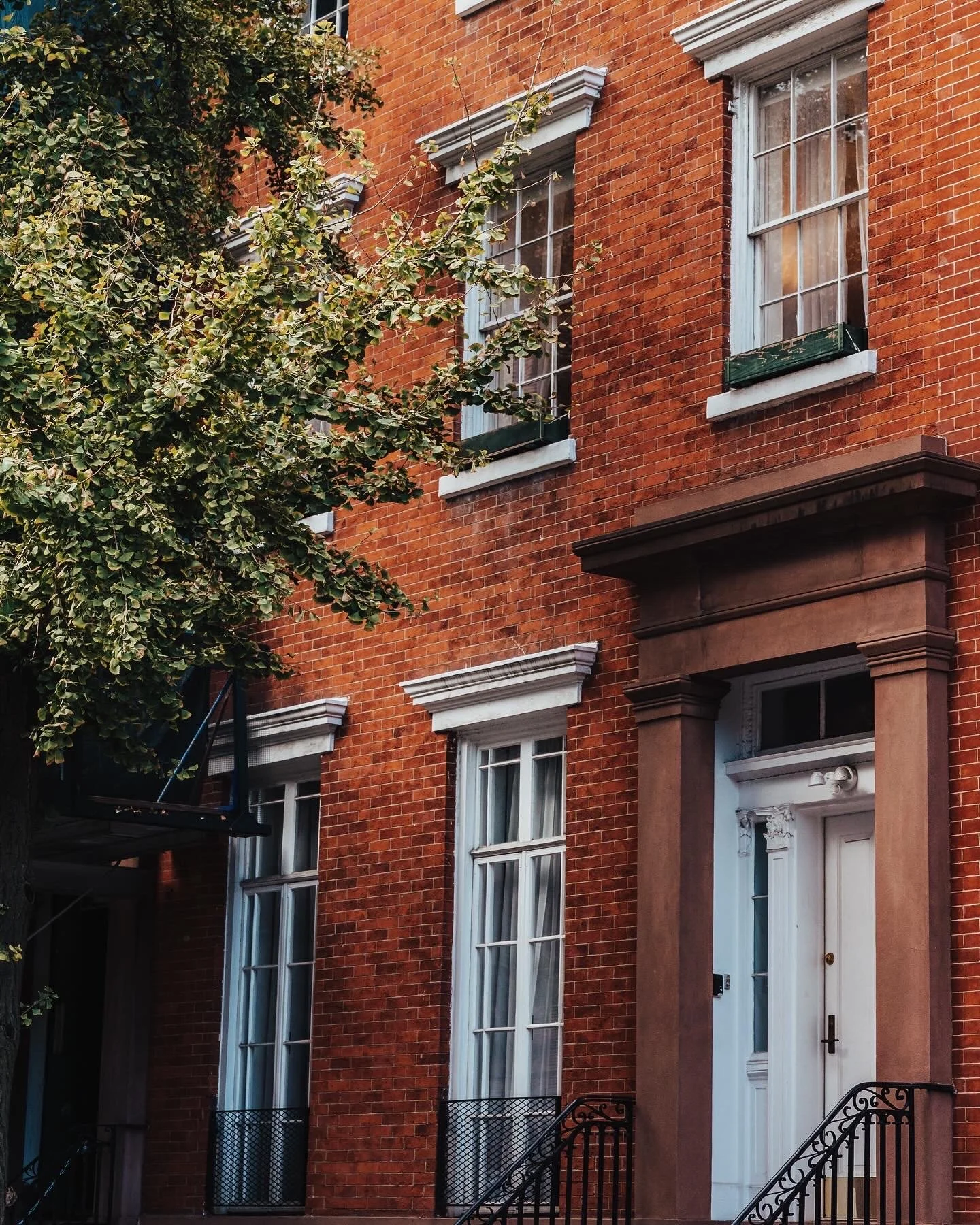 Red brick townhouse with white-framed windows, decorative white trim, and black wrought iron stairs and railings, partially obscured by a leafy tree.