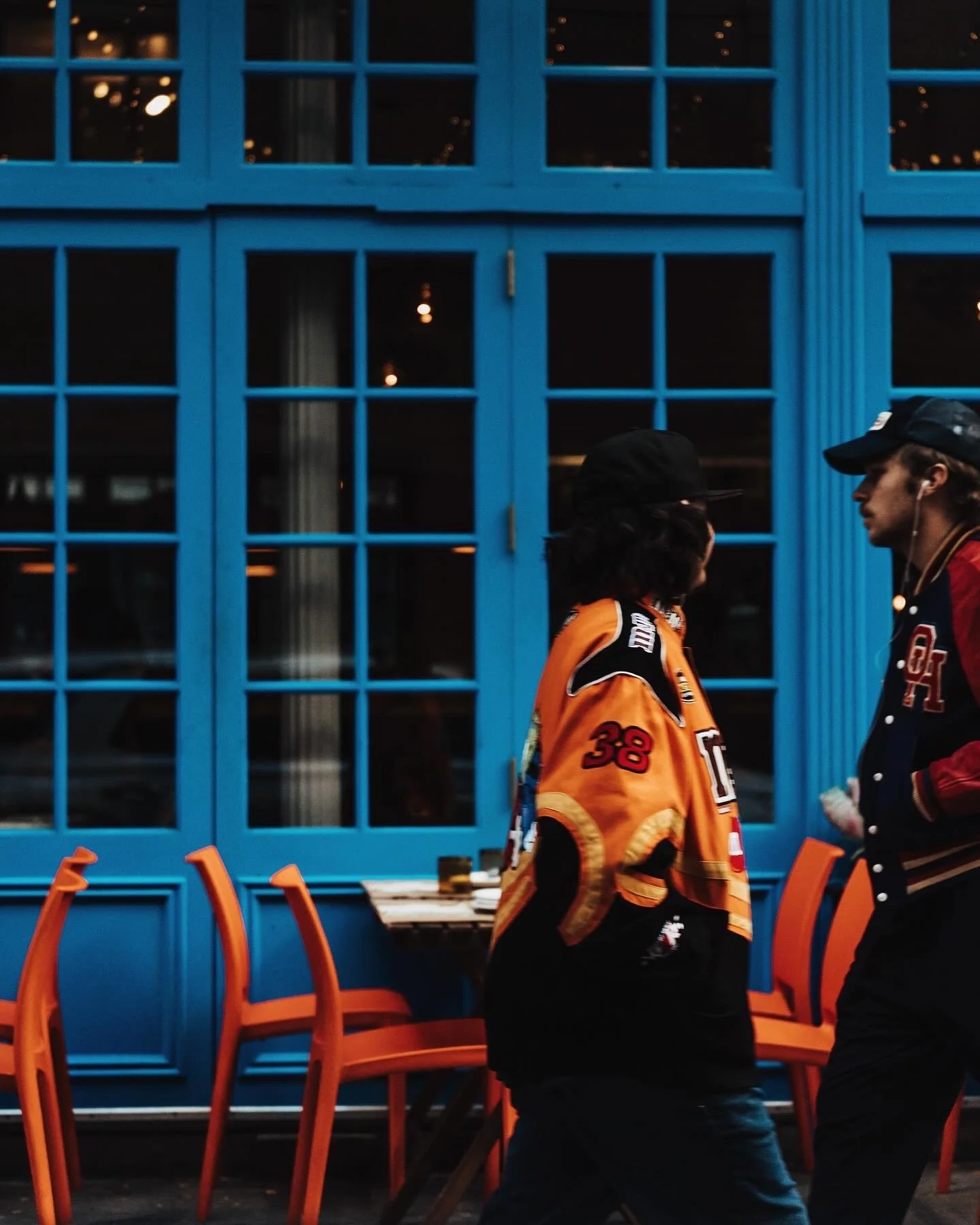 Two people walking past an orange chair and a blue window in a restaurant or café setting.