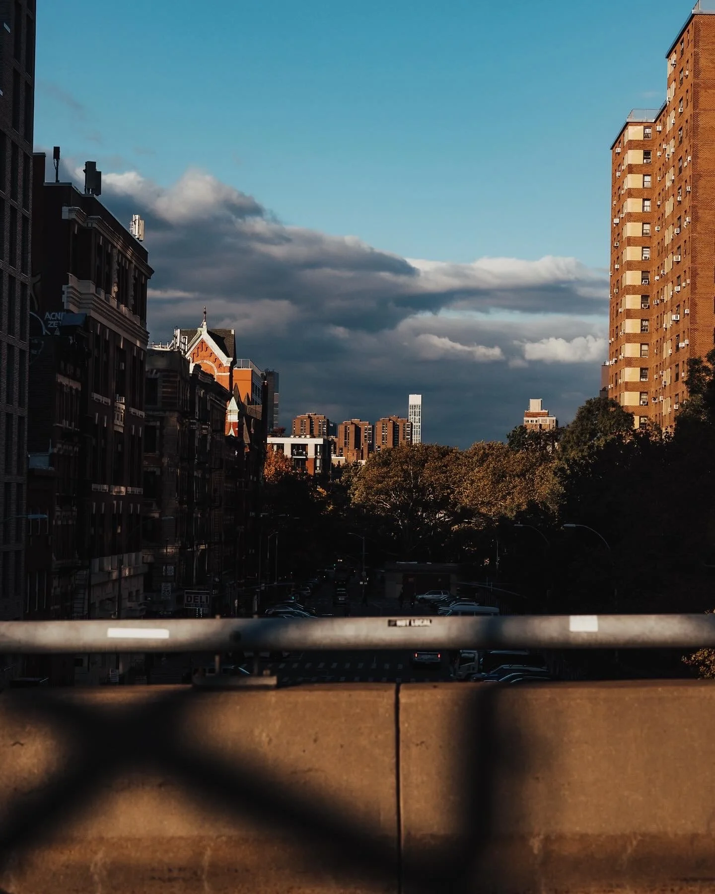 Cityscape view through a chain-link fence, showing buildings, with shadows and clouds in the sky.