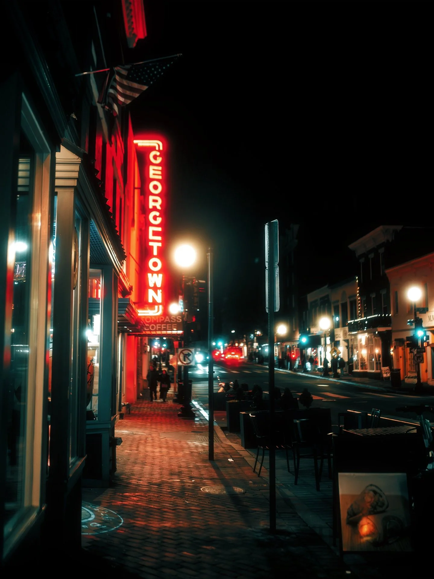 Nighttime street scene with a prominent neon sign reading "GEORGETOWN" and sidewalk cafes. Streetlights cast a glow, and a few pedestrians are visible.