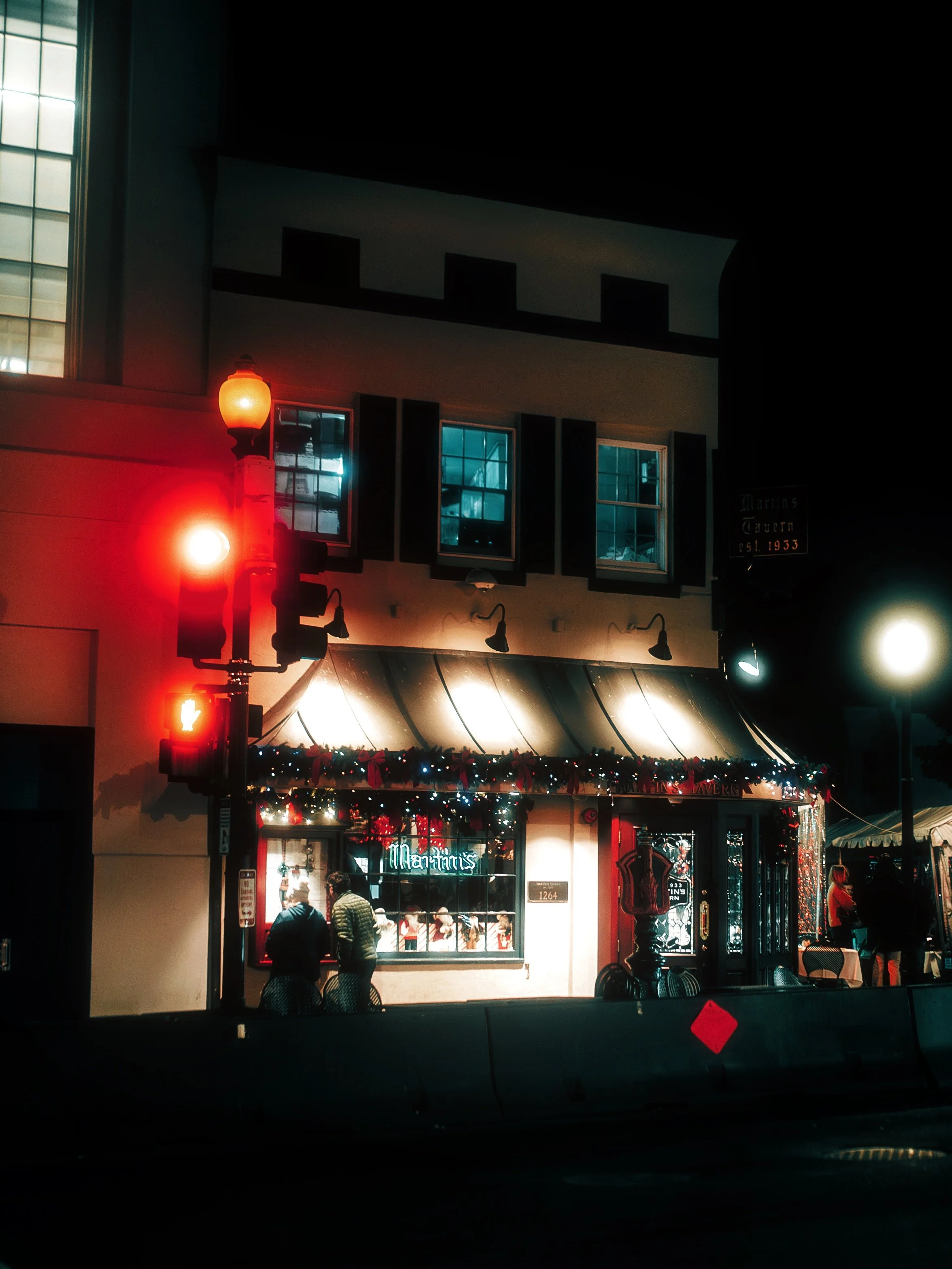 Night scene of a restaurant with holiday decorations, a traffic light showing red and green signals, and a few people near the window.