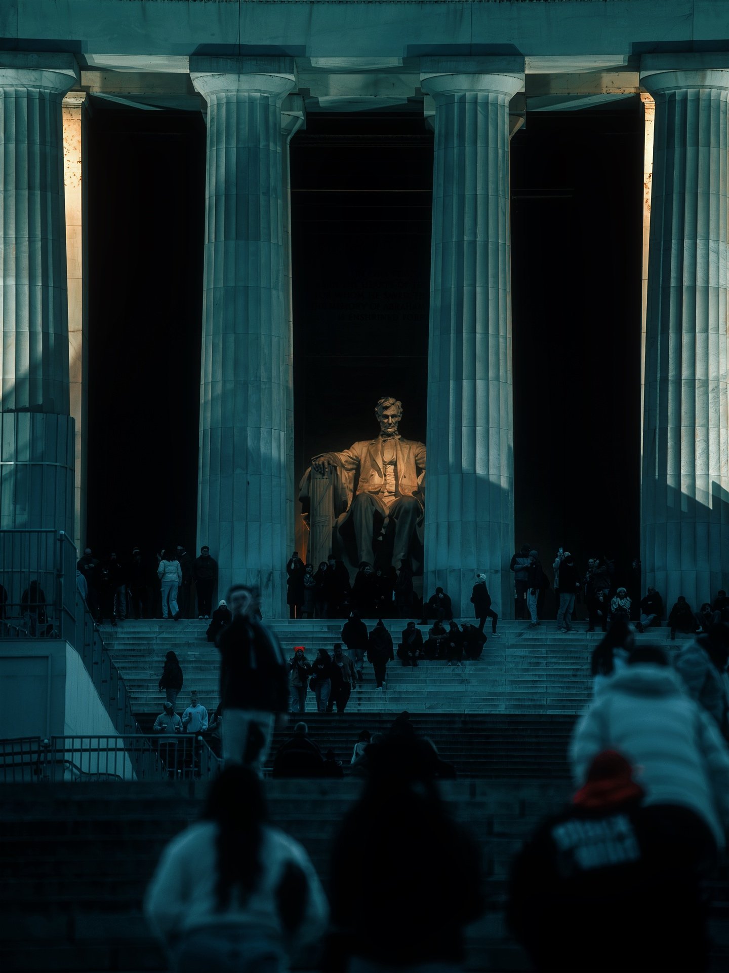 People sitting and standing on the steps in front of the Lincoln Memorial at night, illuminated statue of Abraham Lincoln visible among large columns.