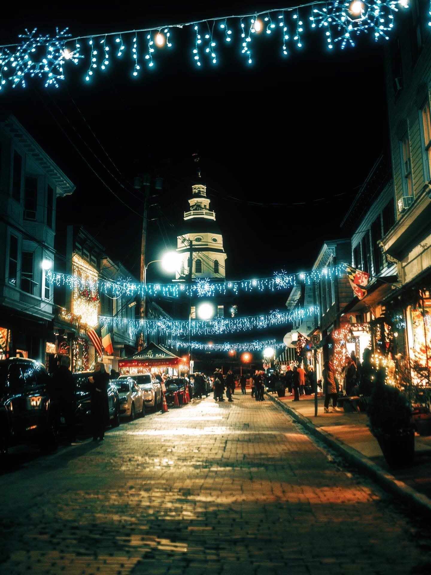 Night view of a decorated street with blue string lights and snowflake ornaments overhead, people walking, cars parked along the sides, and a historic clock tower in the background.