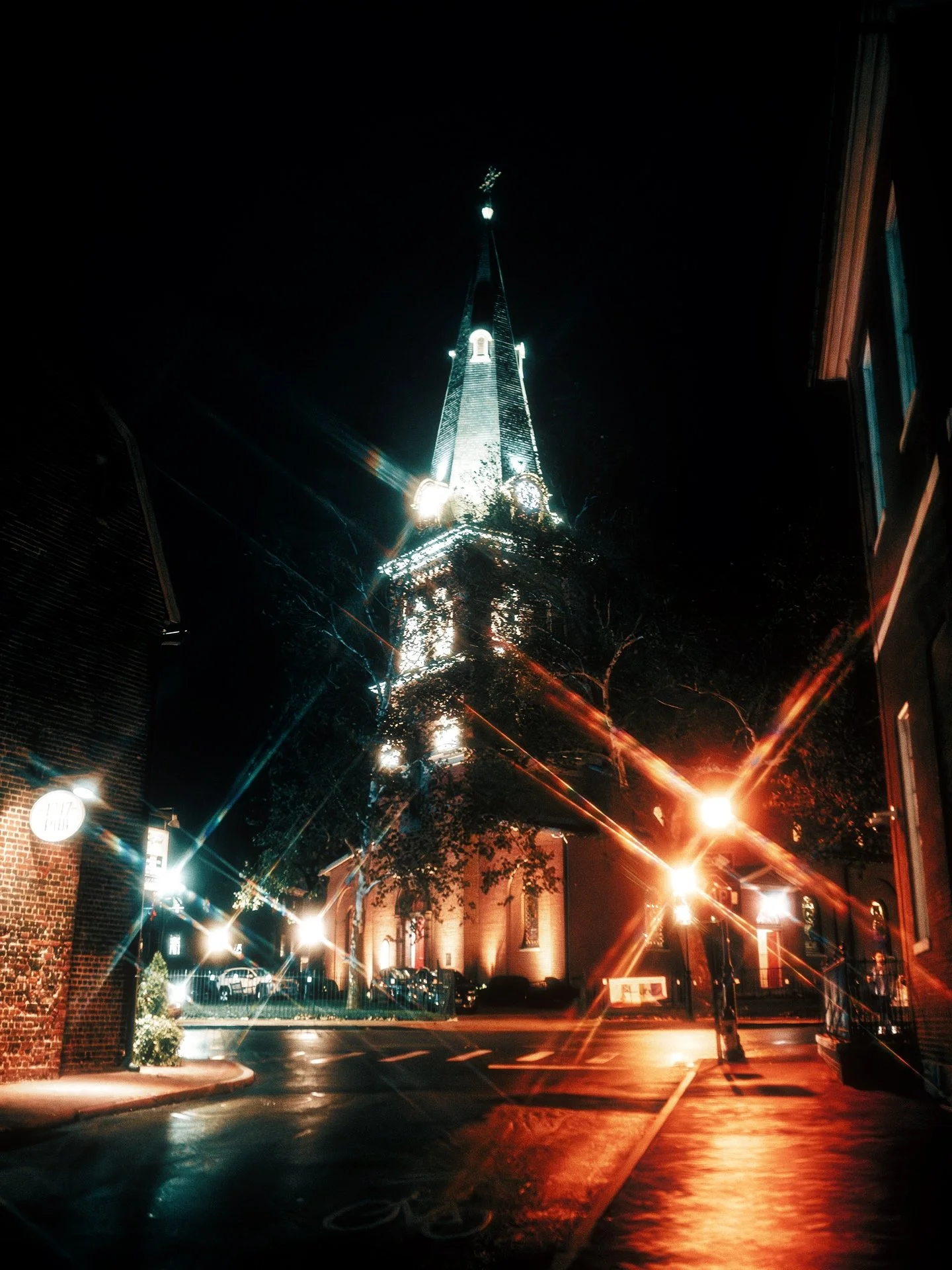 Nighttime view of a church with a tall, illuminated steeple, surrounded by streetlights and buildings.
