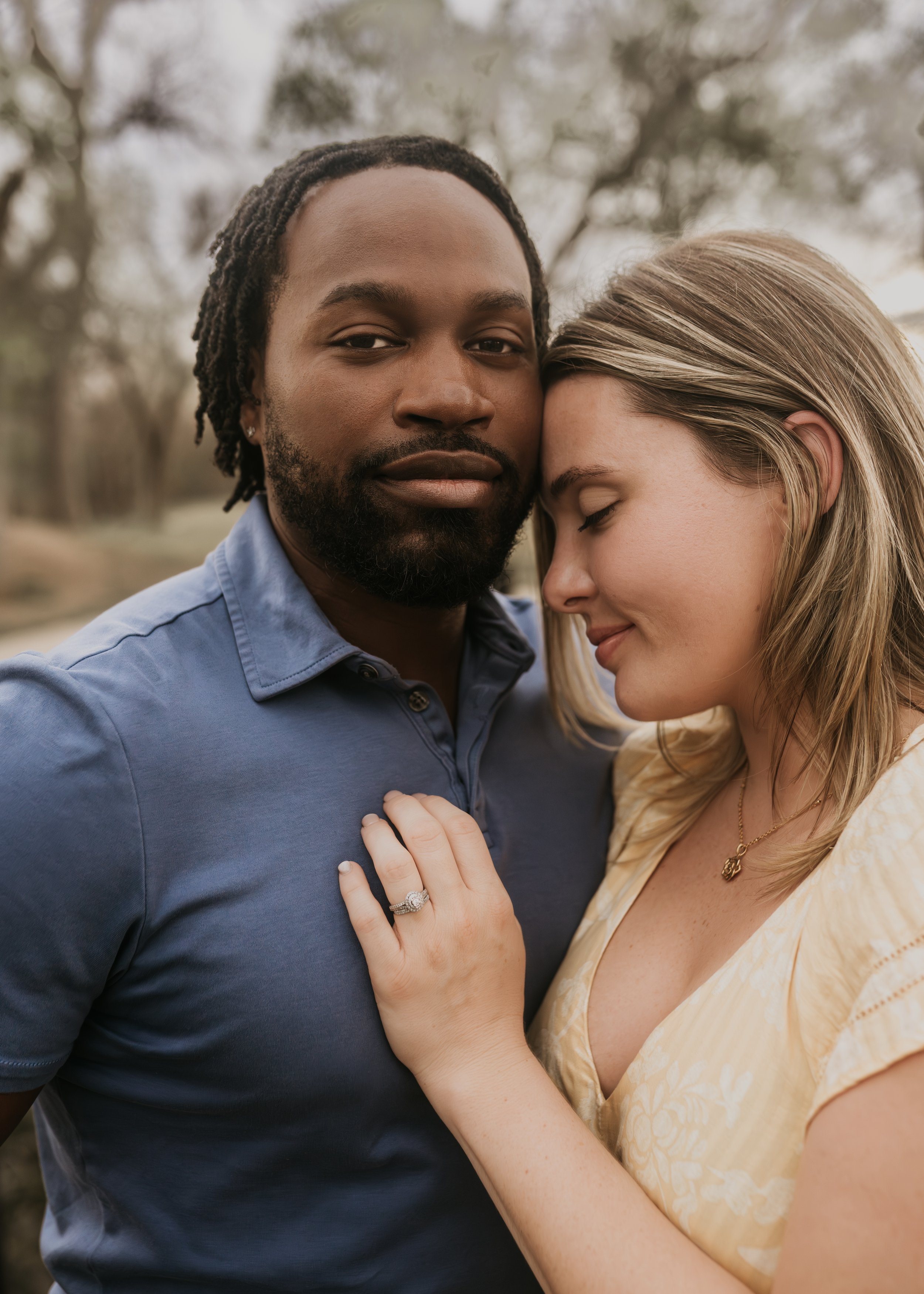 Couple embracing outdoors, woman with engagement ring resting her head on man's chest wearing a blue shirt.