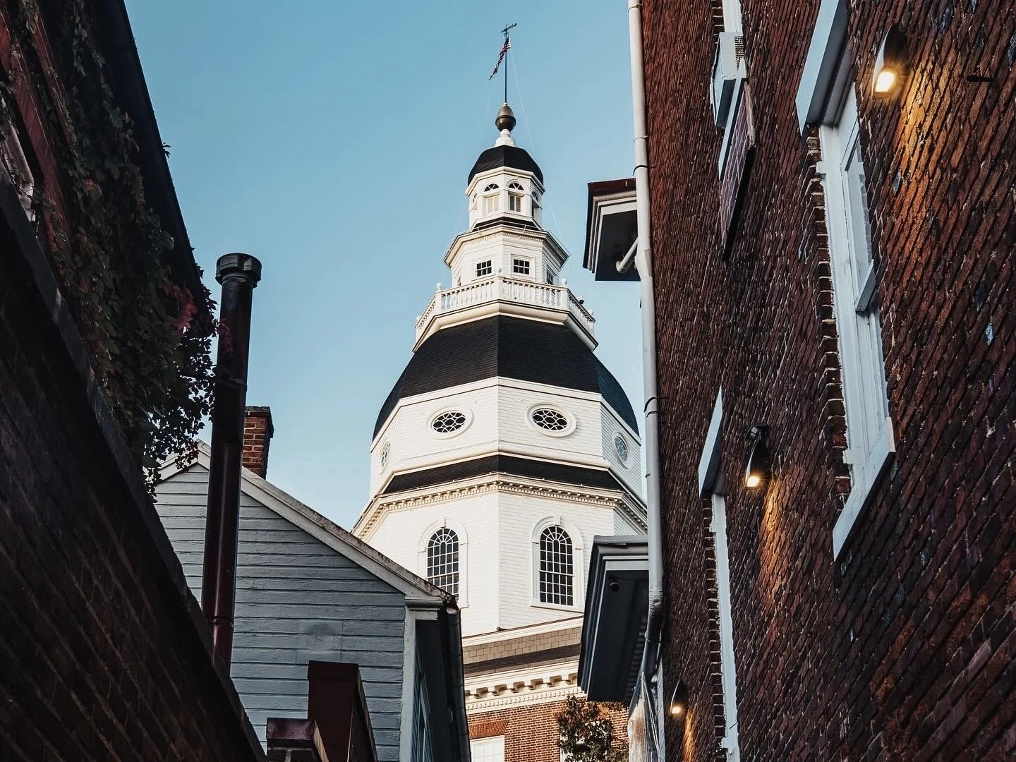 Looking up between narrow alleyways at the State Capitol of Maryland in Annapolis with black accents and a weather vane at the top, flanked by brick and wood-sided buildings with exterior lights.