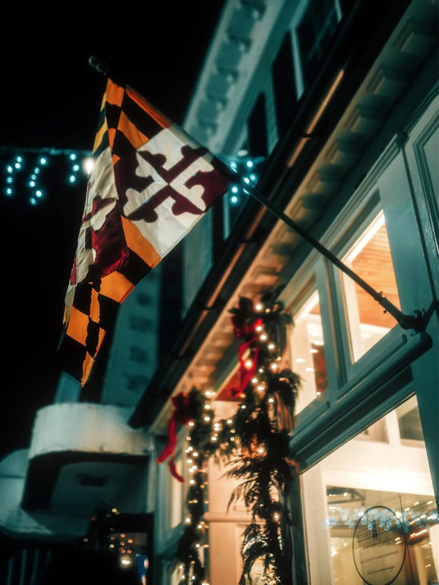A Maryland state flag hanging from a pole outside a decorated building with Christmas garlands and lights.
