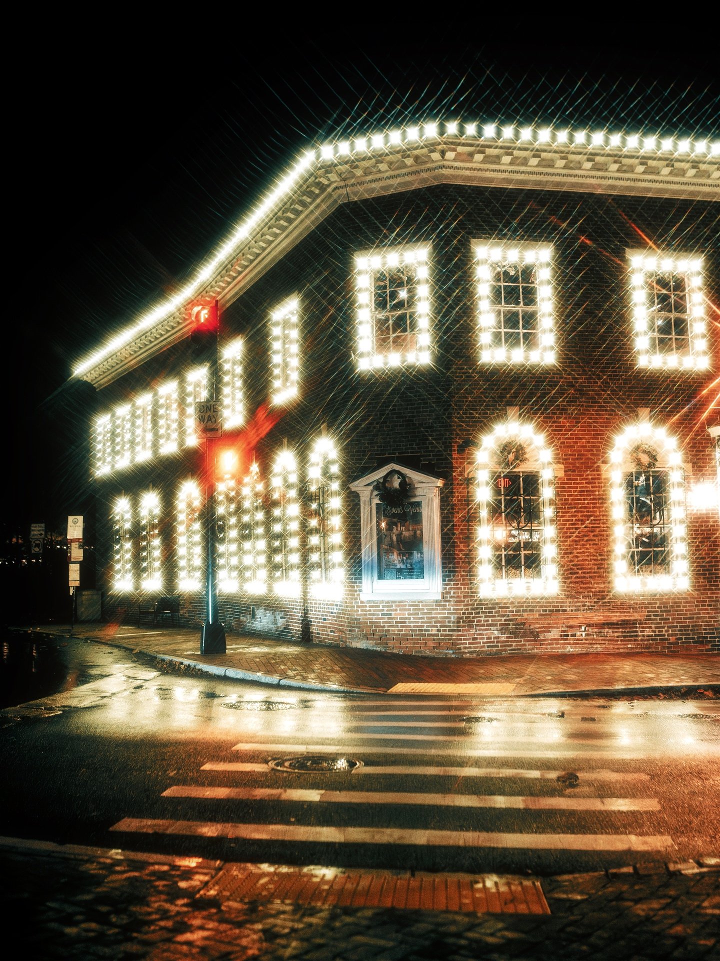 Nighttime street scene with a brick building decorated with white Christmas lights outlining windows and roof, with a crosswalk and wet pavement reflecting the lights.